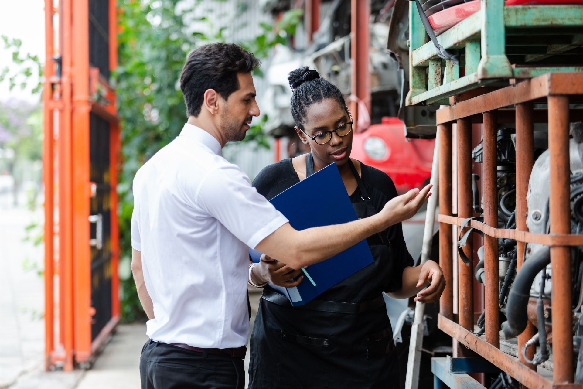 A person in a white shirt and another in a black uniform discuss car parts in a warehouse, holding a blue folder.