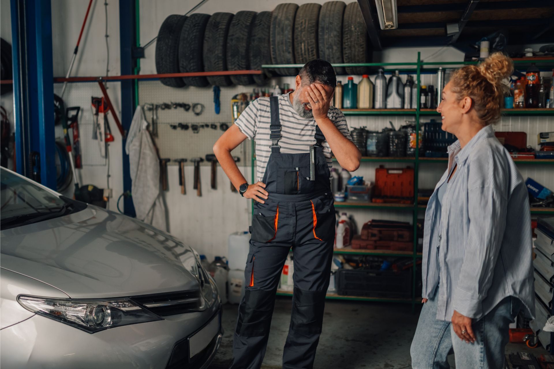 A mechanic in overalls covers their face in frustration while speaking to a customer in a car repair shop.