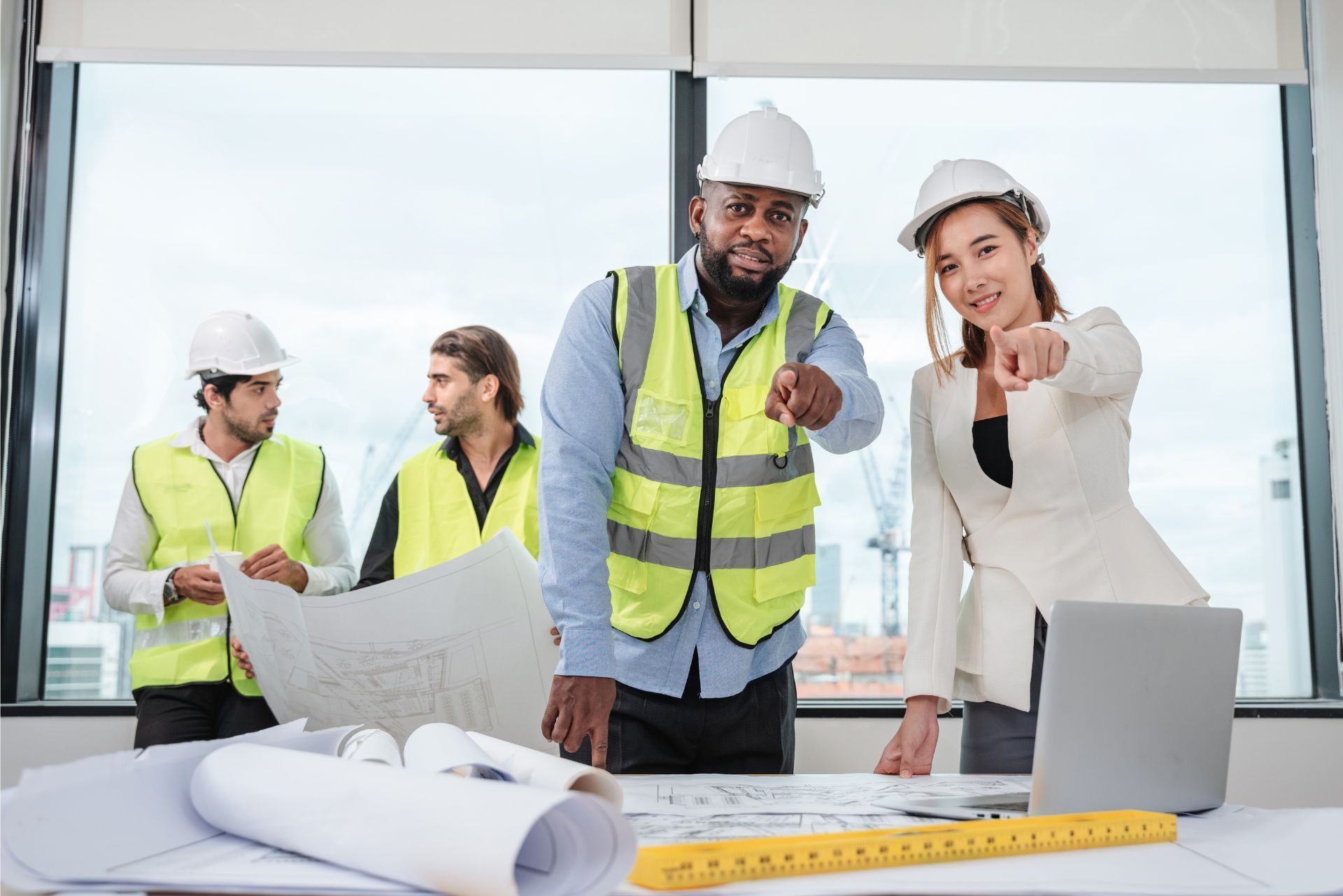 Professionals in safety gear and office attire reviewing blueprints in a sunlit office, pointing toward the viewer.