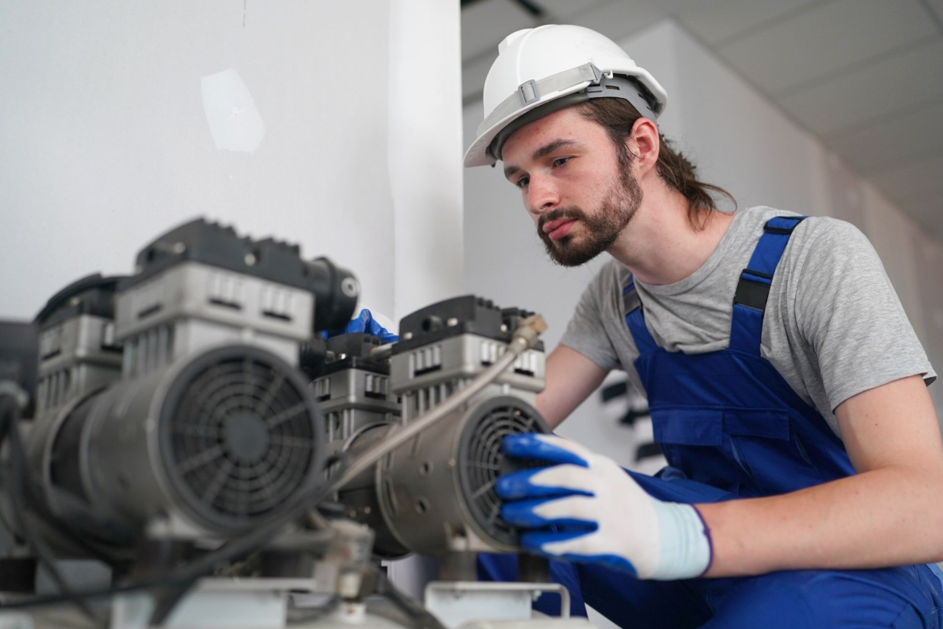 Technician in white hard hat and blue overalls inspecting industrial machinery in a workshop