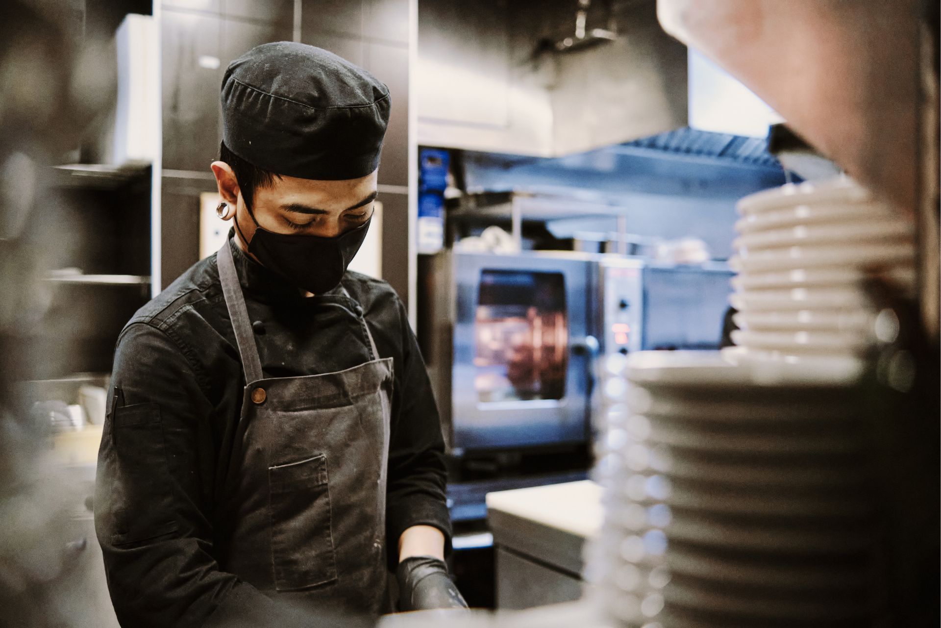 A chef in black uniform, apron, cap, and mask focuses on food preparation in a commercial kitchen with stacked plates.