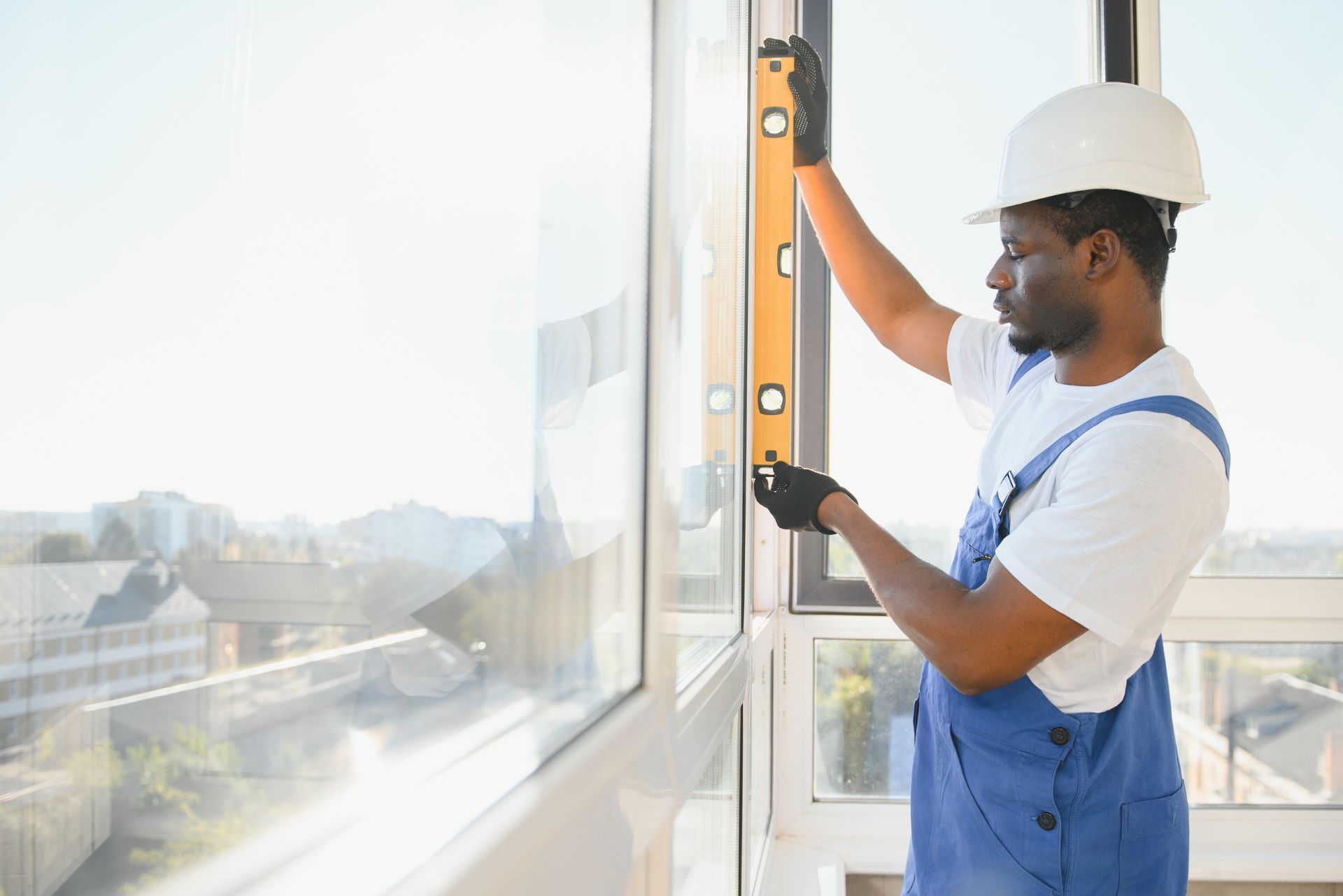 Worker in a white hard hat measures a window with a level in a bright room.