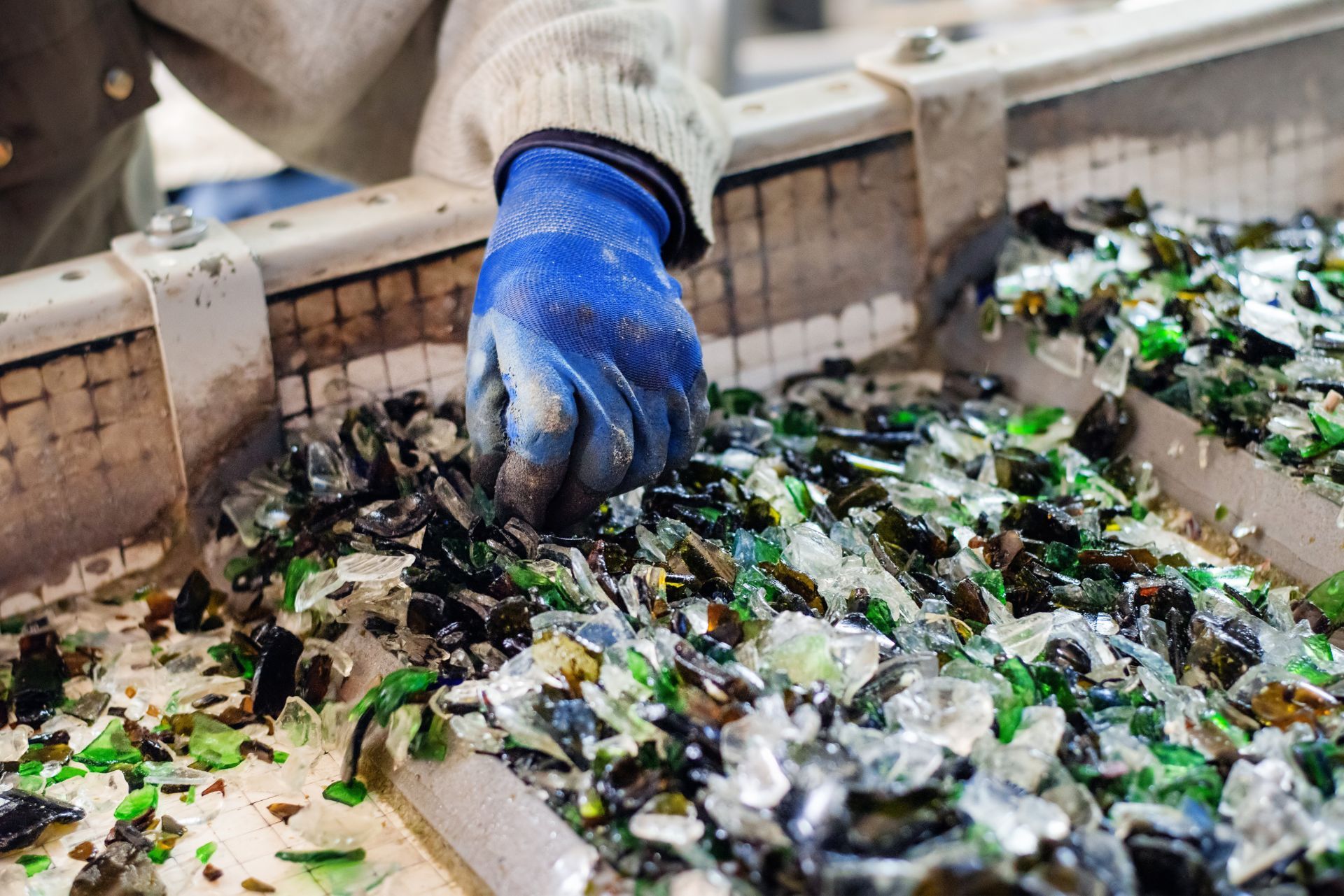 A gloved hand sorts through a conveyor belt filled with mixed pieces of crushed glass at a recycling facility.
