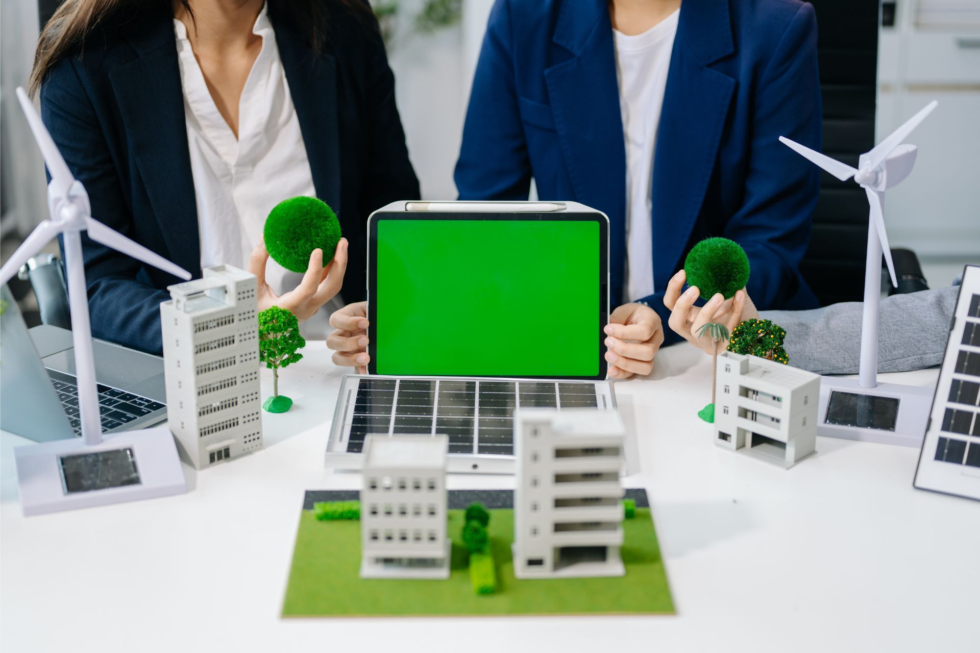 Two people hold green spheres behind a laptop with a green screen, surrounded by models of buildings and wind turbines.