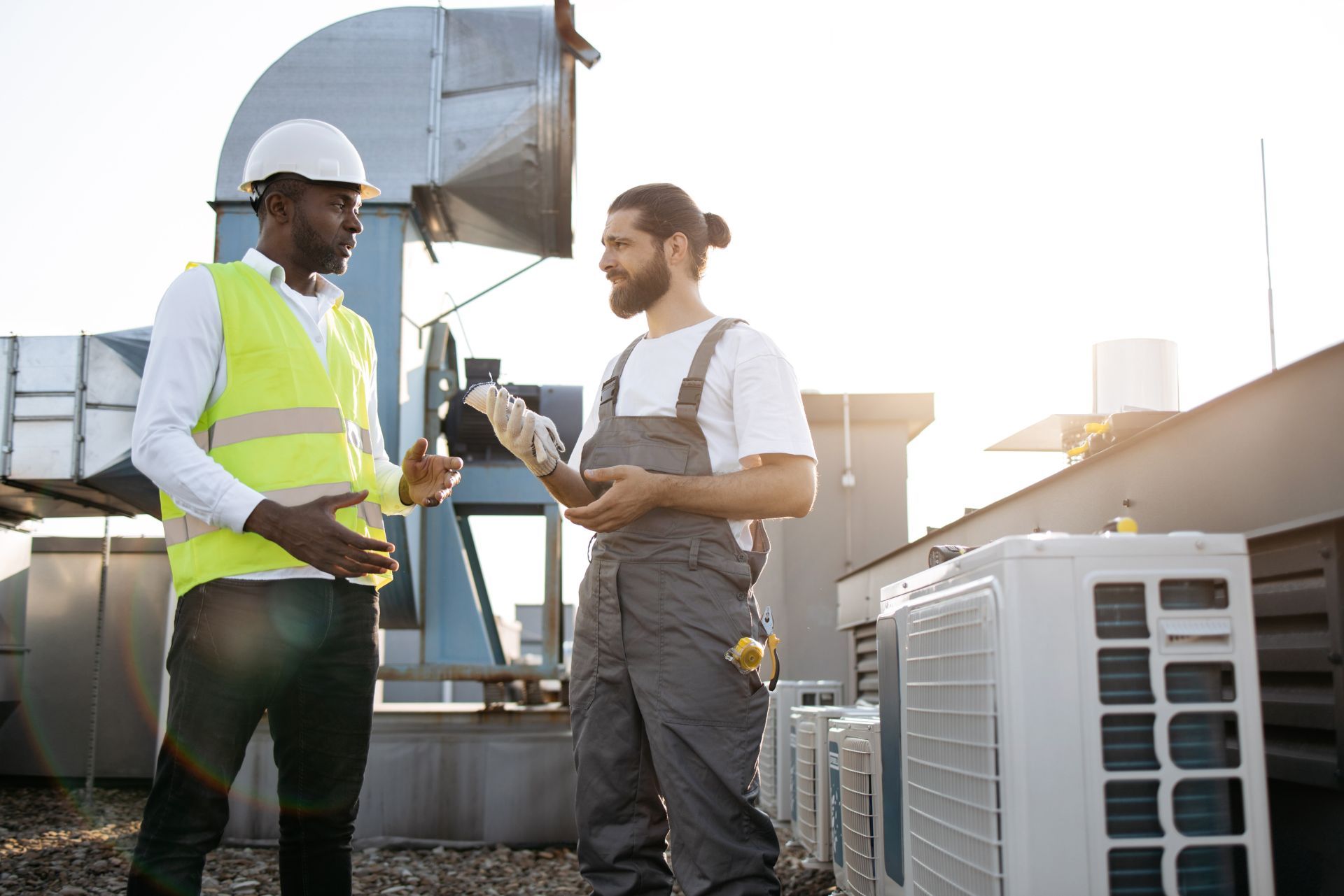 Two workers in safety gear talking beside rooftop HVAC units in bright sunlight