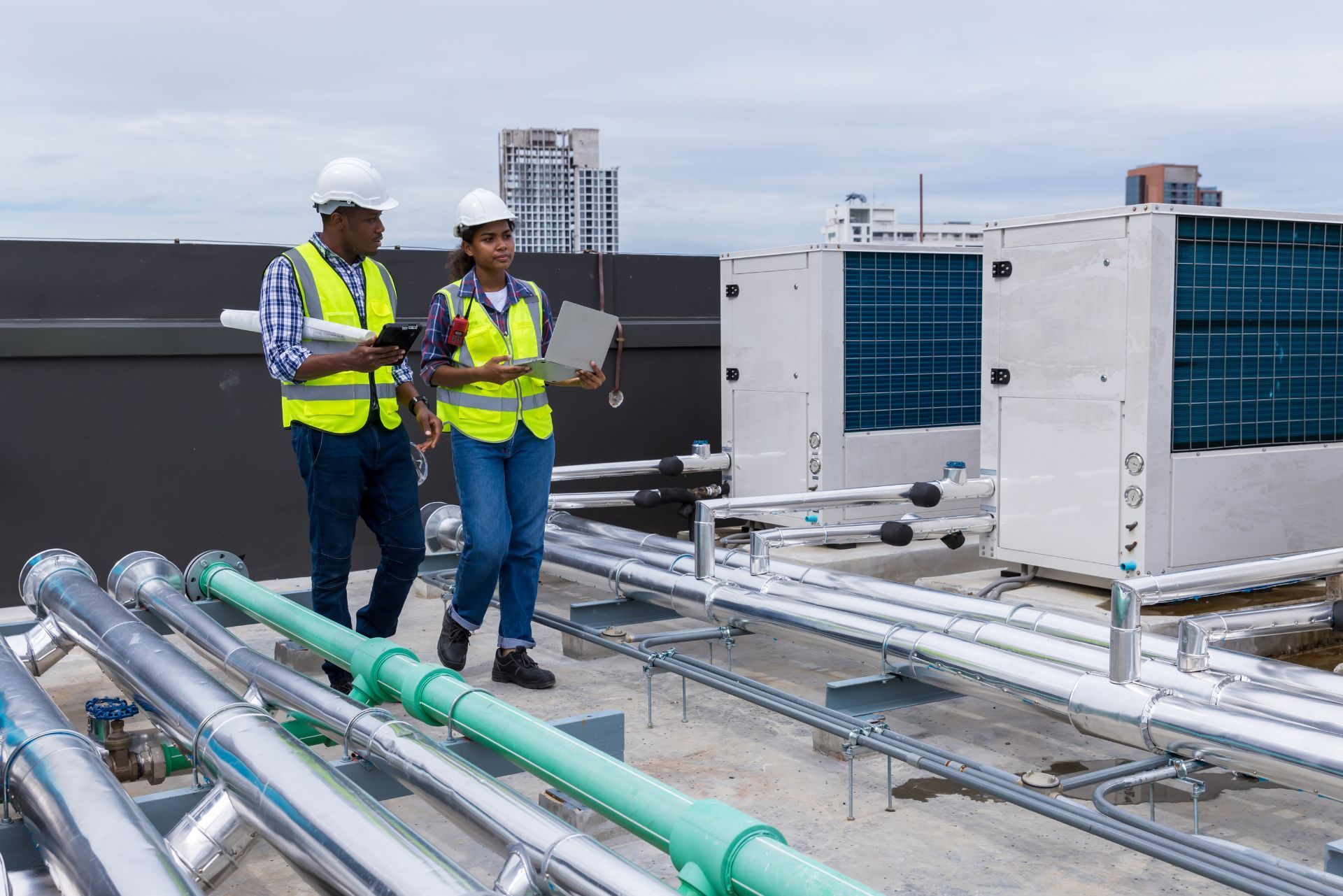 Two engineers in safety vests inspect rooftop HVAC units and pipes