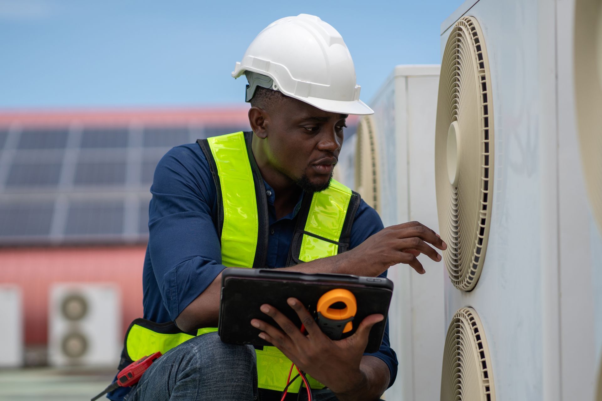 Technician in hard hat and safety vest using a tablet beside rooftop HVAC equipment.