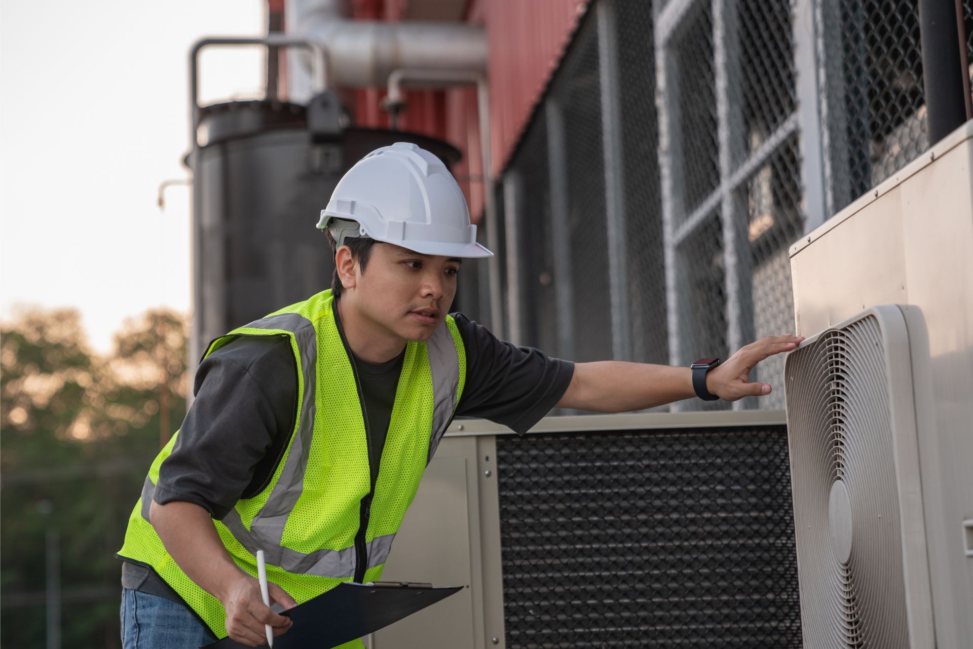Worker in a hard hat and safety vest inspecting an outdoor HVAC unit.