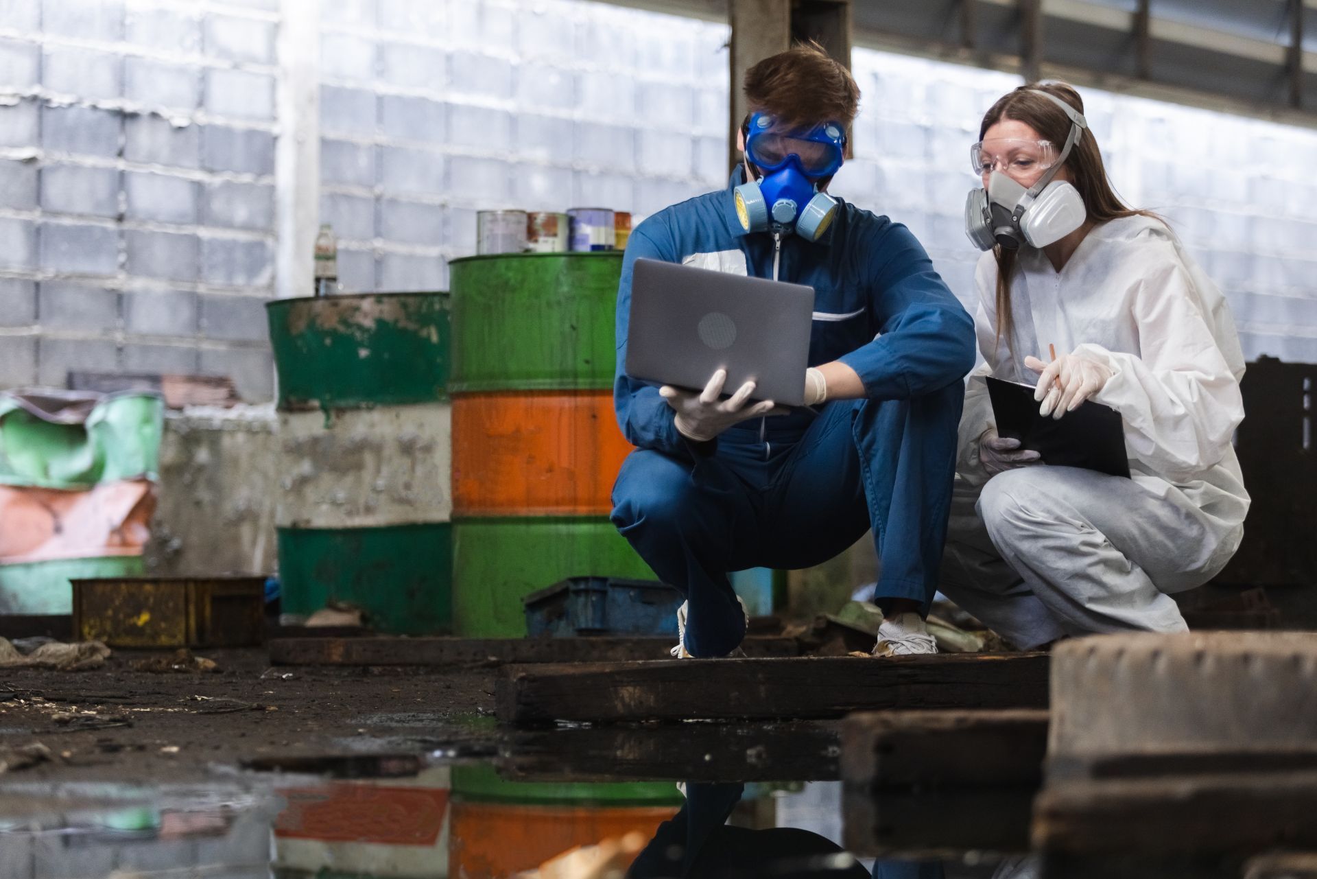 Two people in hazmat gear and respirators crouch in an industrial warehouse, examining data on a laptop and clipboard.