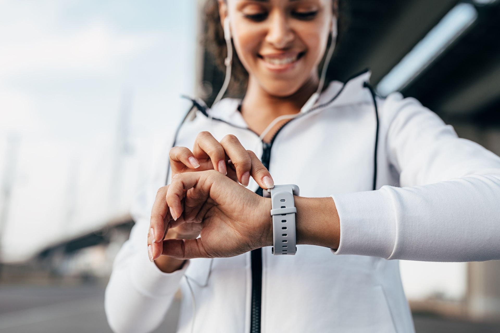 A person in a white workout jacket checks their grey smartwatch while wearing earbuds outdoors.