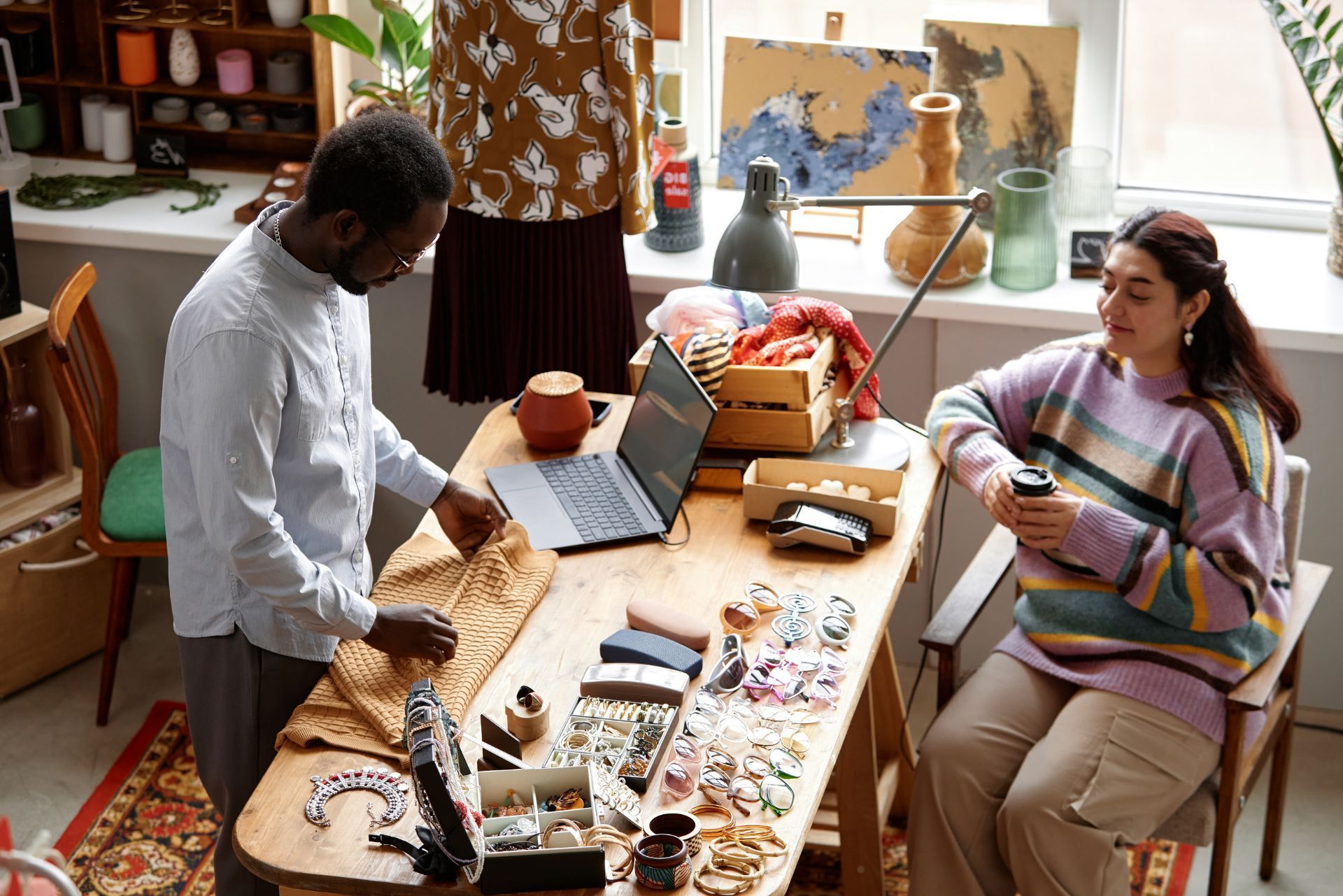 Two people working at a craft table in a bright studio, surrounded by paper, tools, and art supplies.