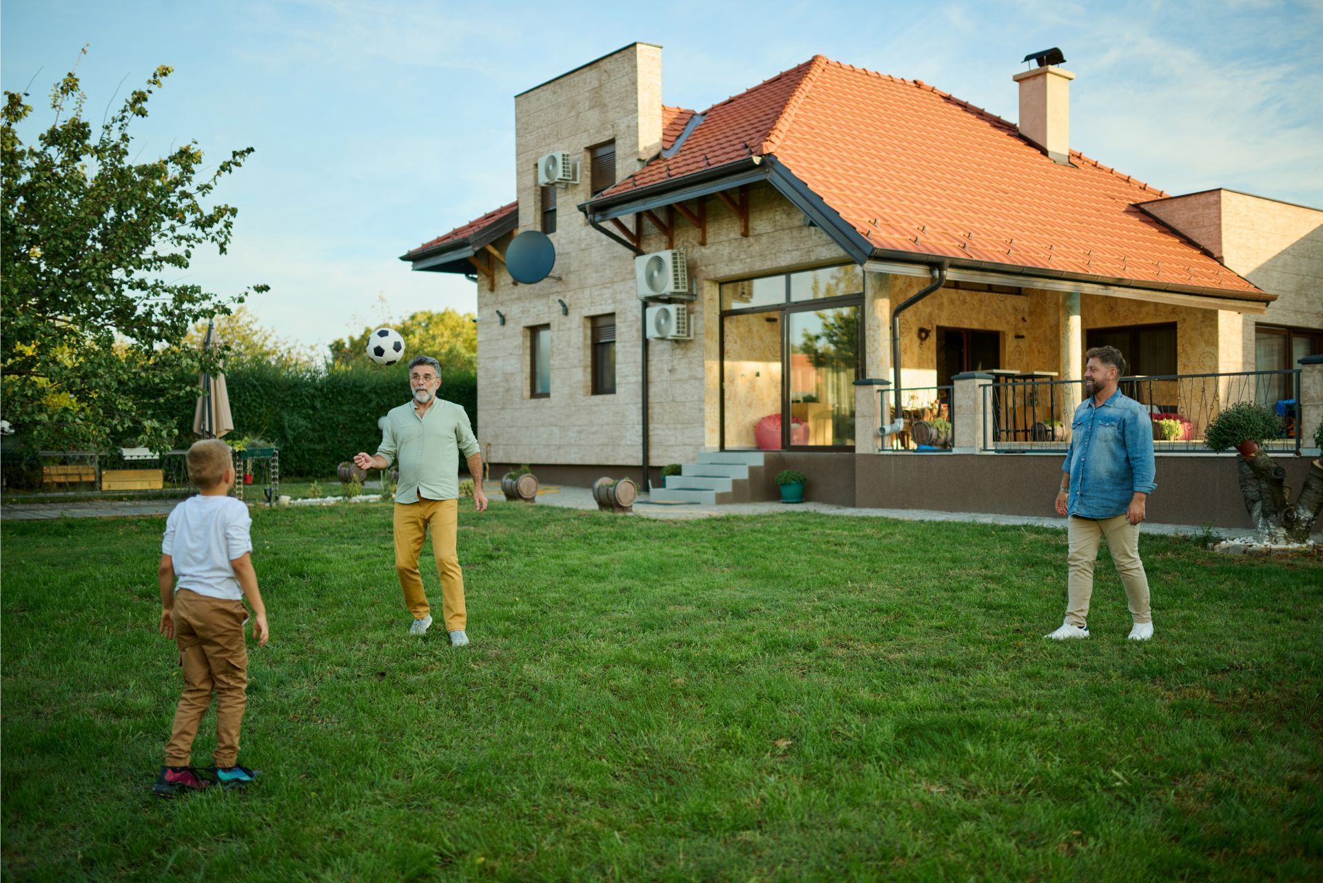 Three people standing on a lawn in front of a modern house with a red roof and stone exterior.