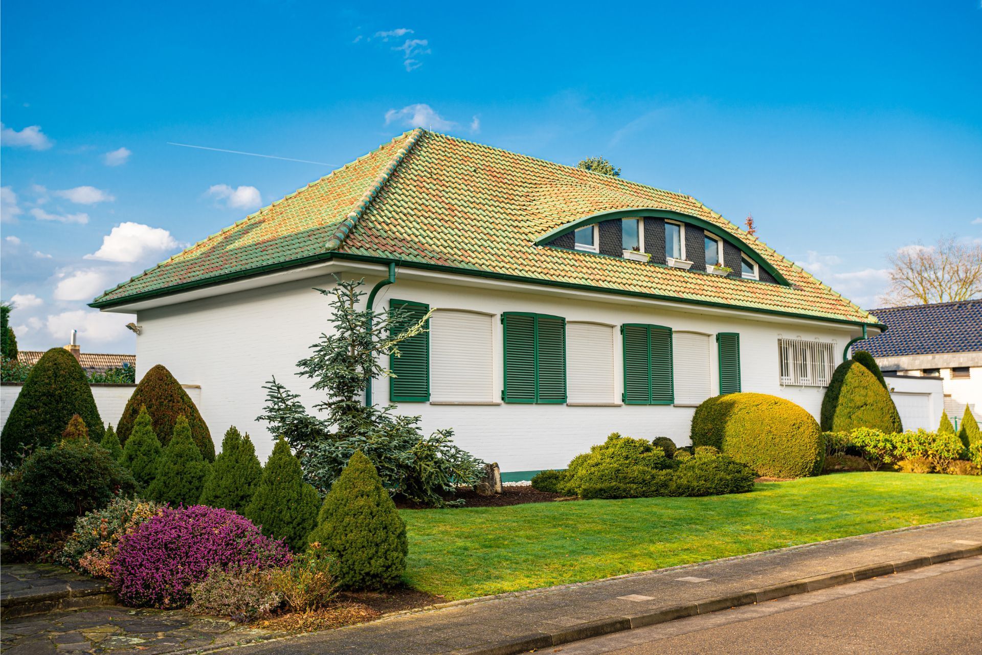 Single-story house with green shutters, tiled roof, and manicured garden under a blue sky