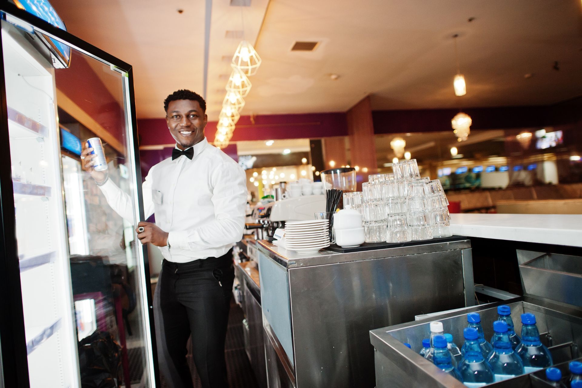 Waiter in white shirt and black bow tie stands by a drink station in a banquet hall, smiling.