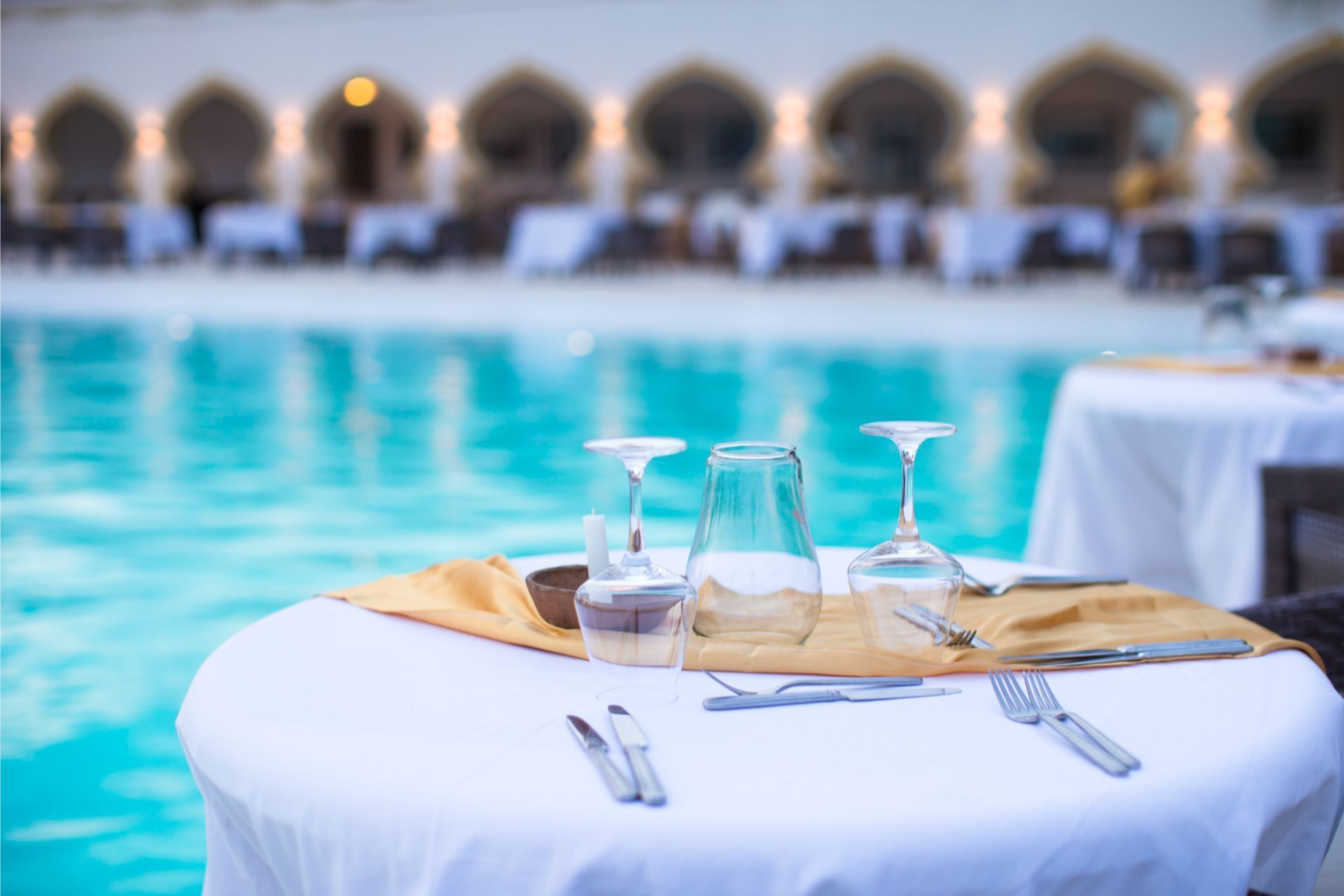 Table set beside a turquoise pool with glasses, cutlery, and a folded napkin at an outdoor dining area
