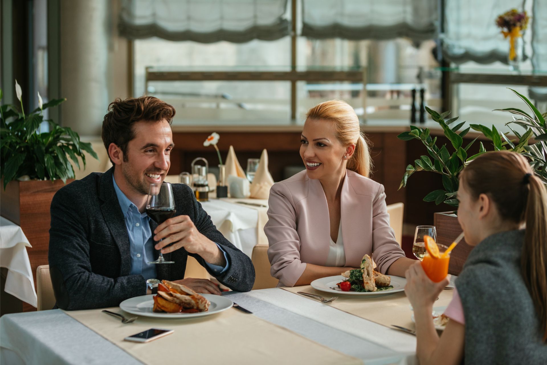 Three people chatting over lunch at a bright restaurant table, smiling and holding drinks.
