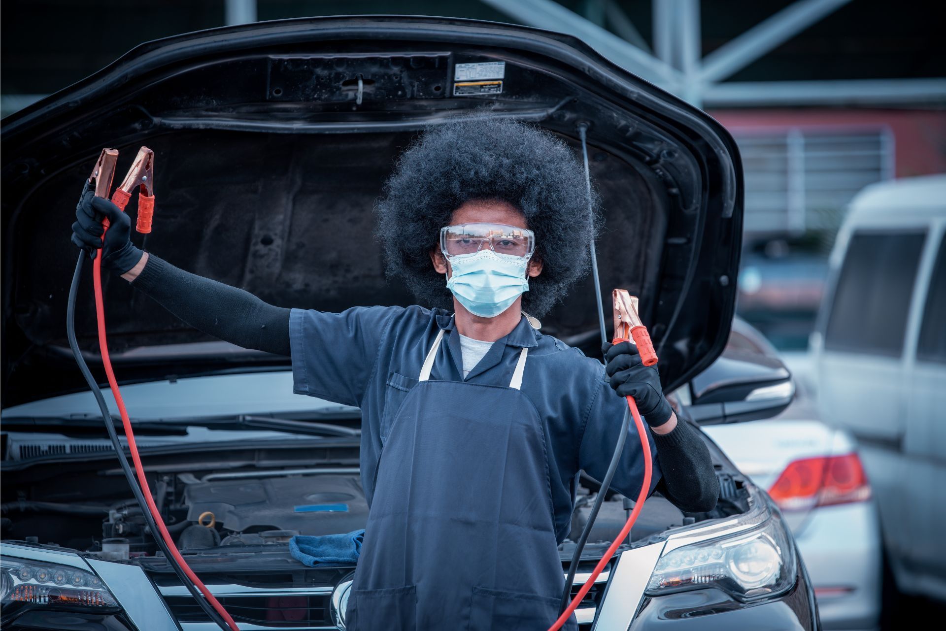 Mechanic wearing a mask holding jumper cables in front of an open car hood