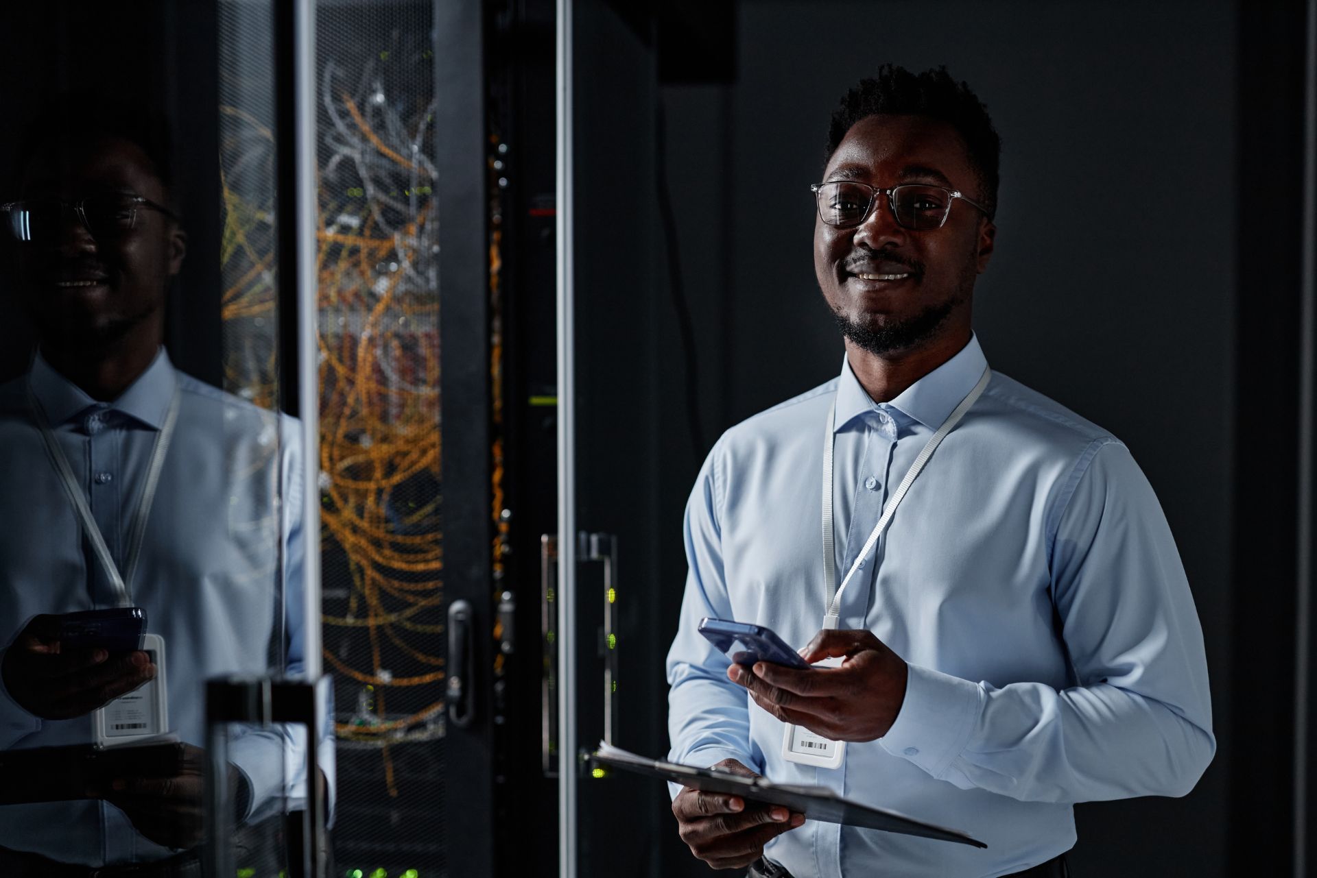 Man in a white shirt and tie standing in a server room, holding a phone and tablet, with reflected racks beside him
