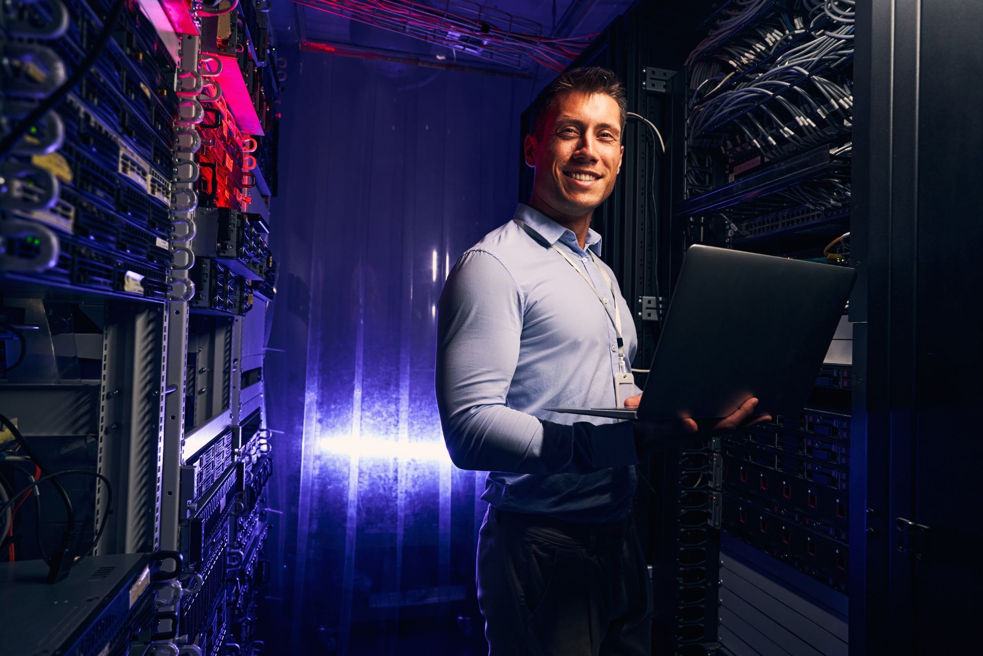 Technician in a server room holding a laptop, surrounded by glowing rack servers and purple lights