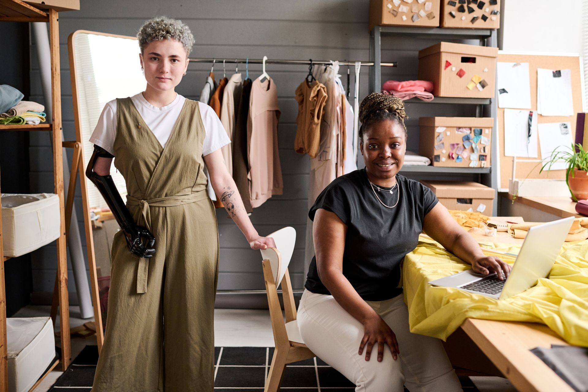 Two people stand in a design studio; one with a prosthetic arm smiles, and the other sits at a desk using a laptop.