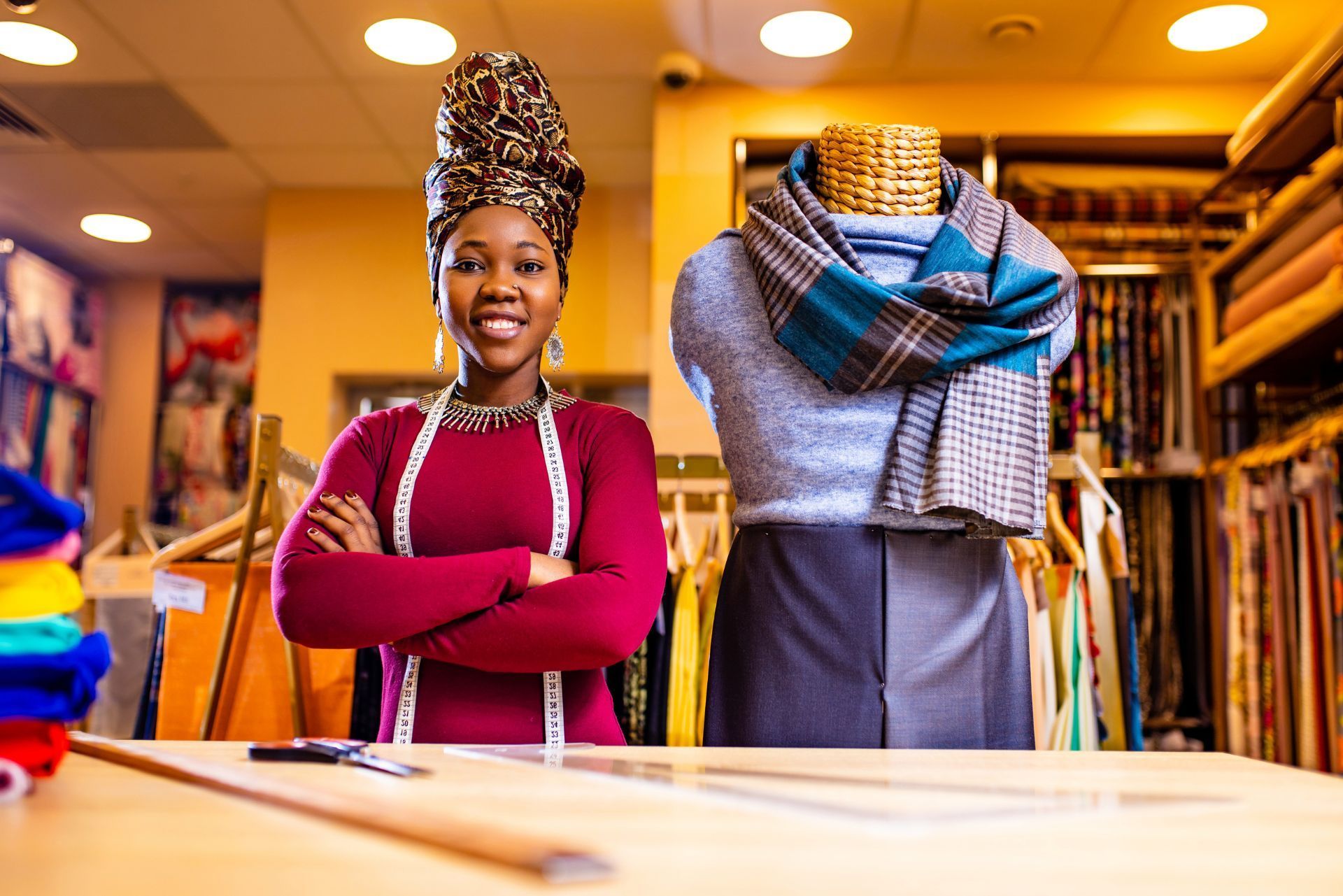 A tailor with a measuring tape stands with arms crossed in a fabric store next to a mannequin wearing a scarf.