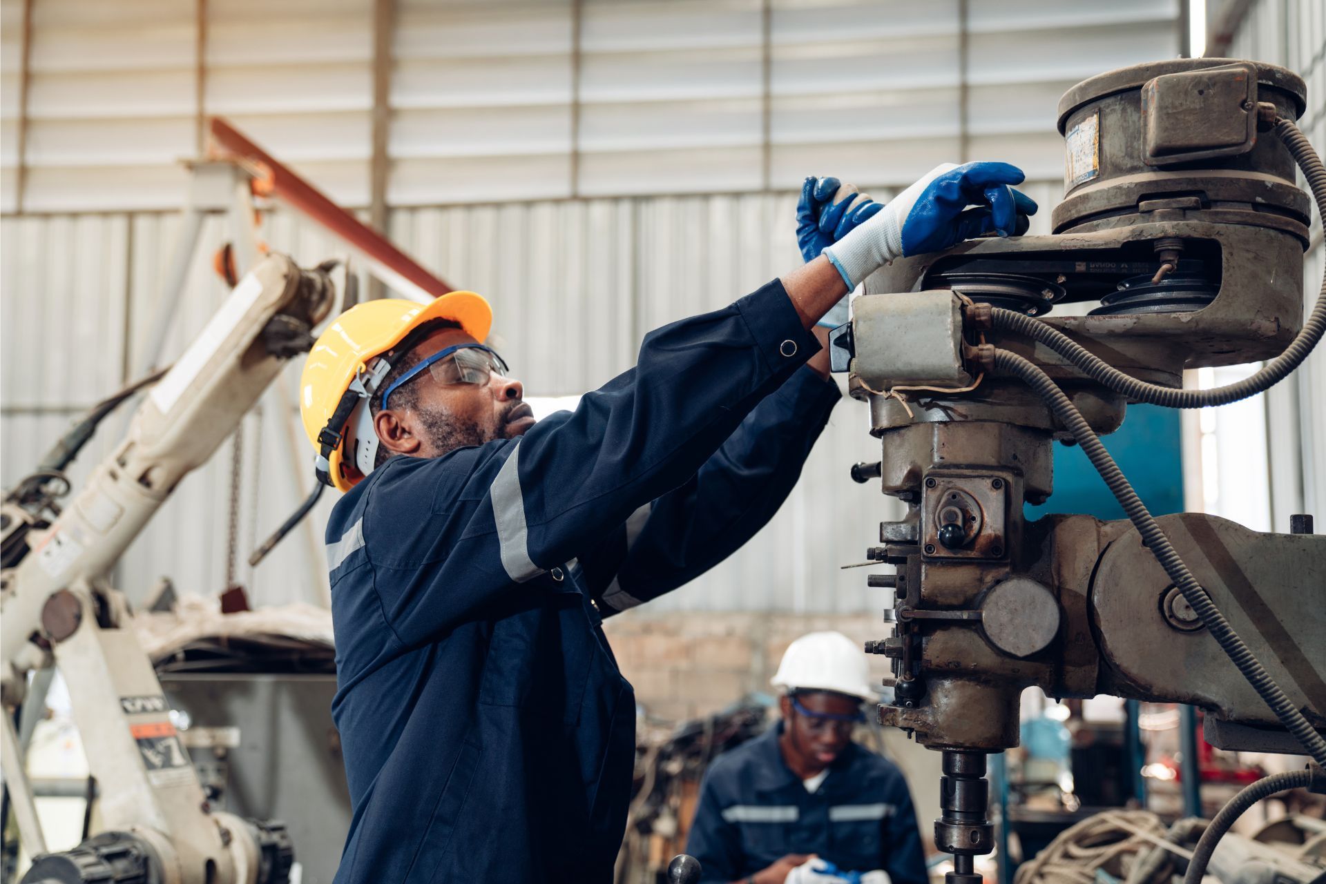 Worker in blue coveralls and yellow hard hat adjusting machinery in an industrial workshop