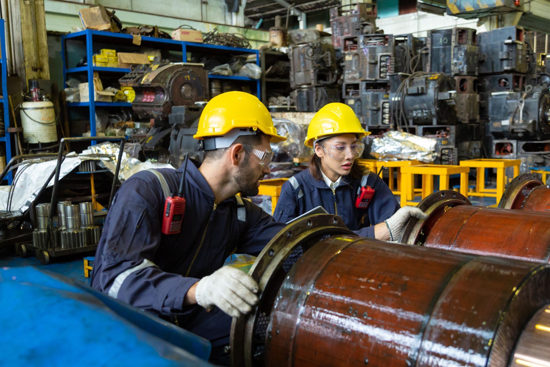 Two workers in yellow hard hats inspecting large metal machinery in a factory workshop