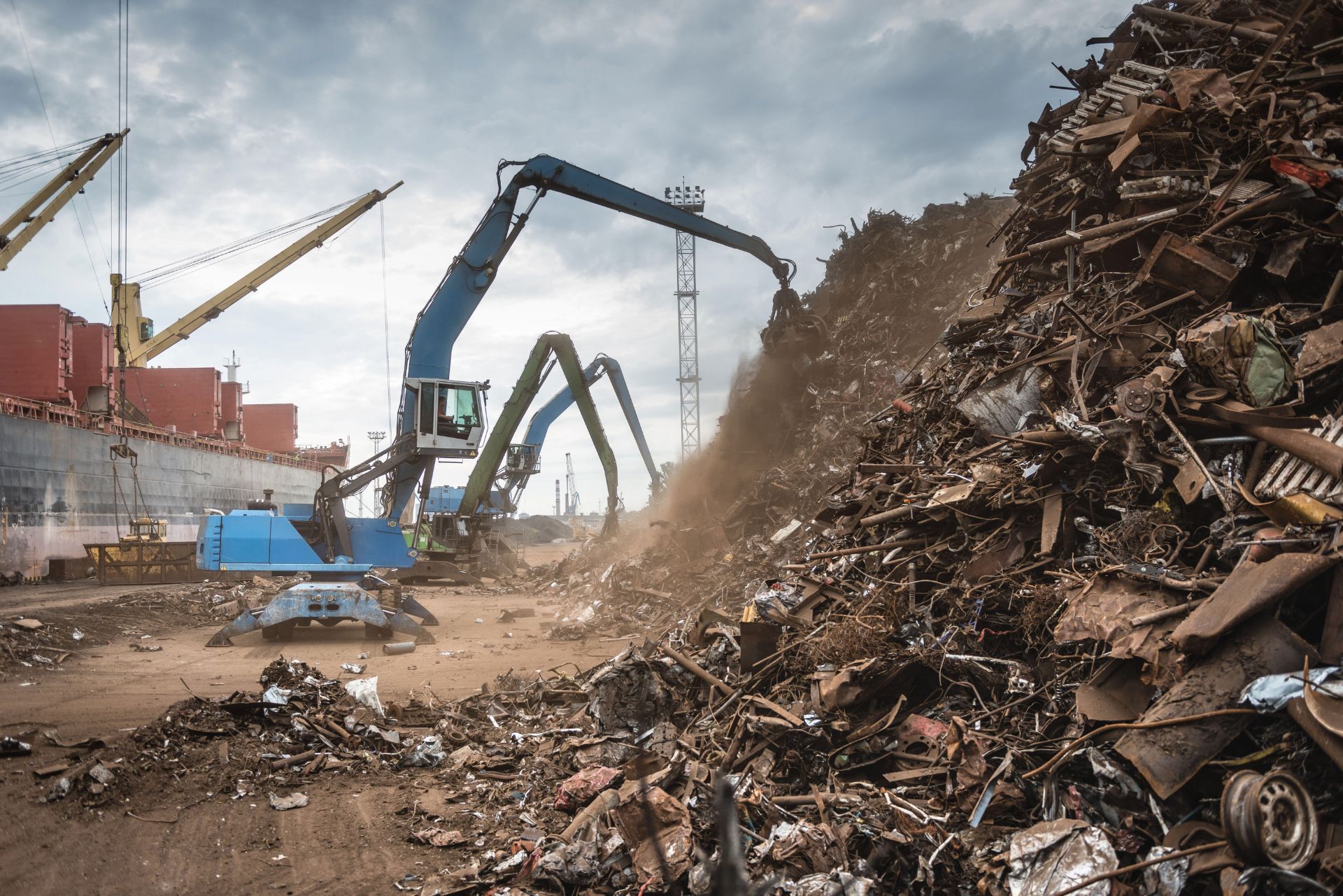 Construction machinery loading a large scrap metal pile at a recycling yard under a cloudy sky