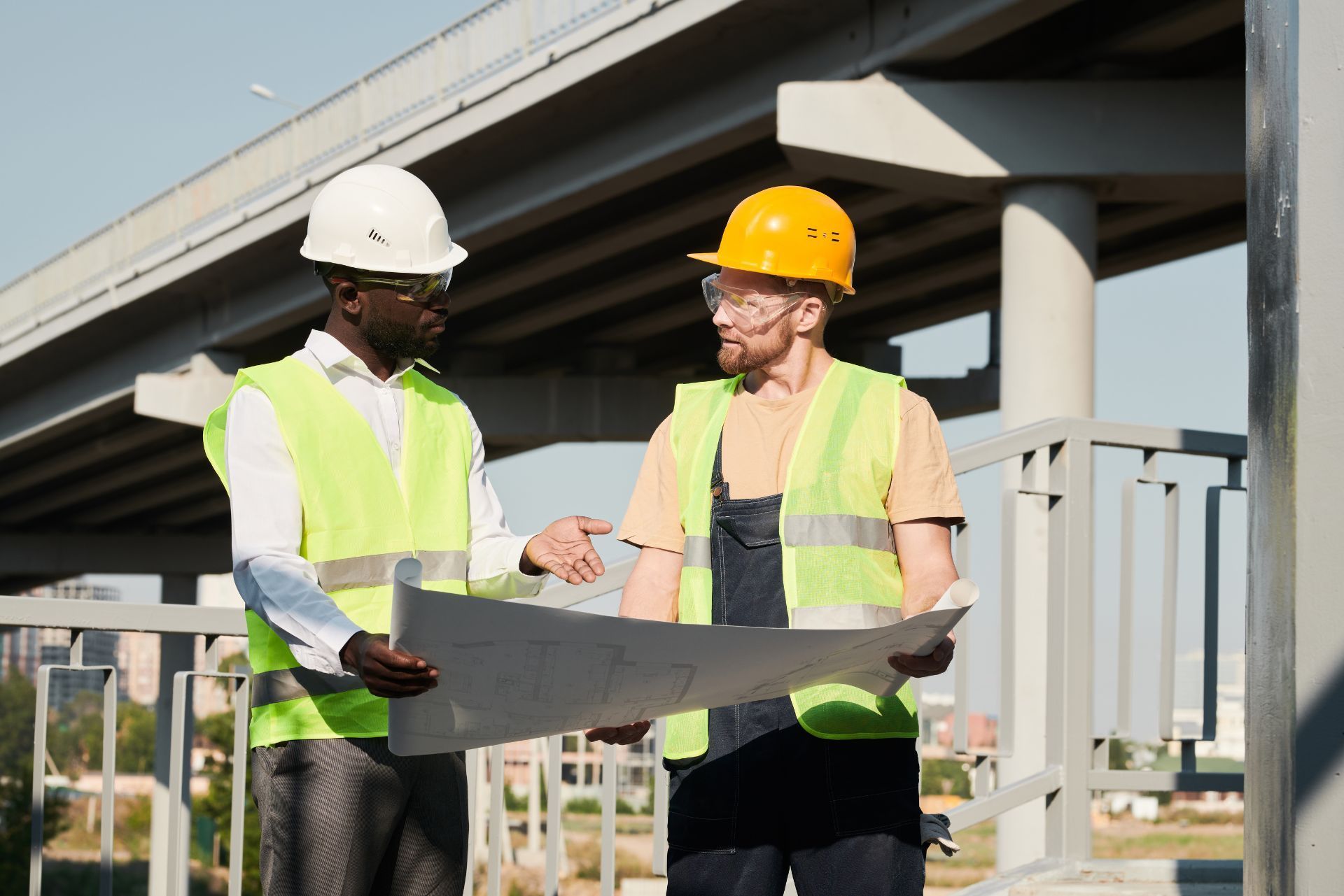 Two construction workers in hard hats reviewing blueprints under an overpass