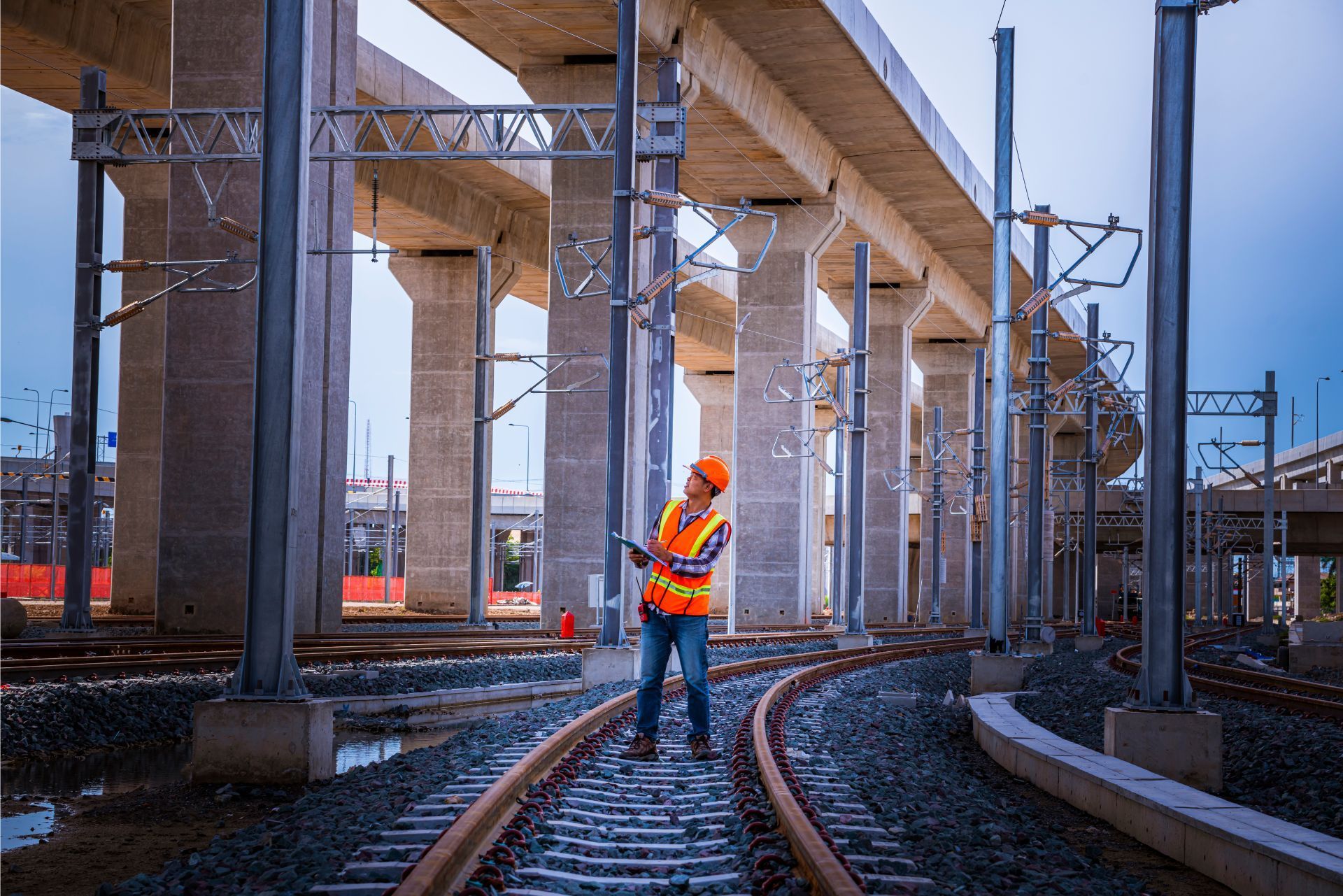 Worker in orange safety gear on railway tracks beneath an elevated bridge, with construction cranes nearby.