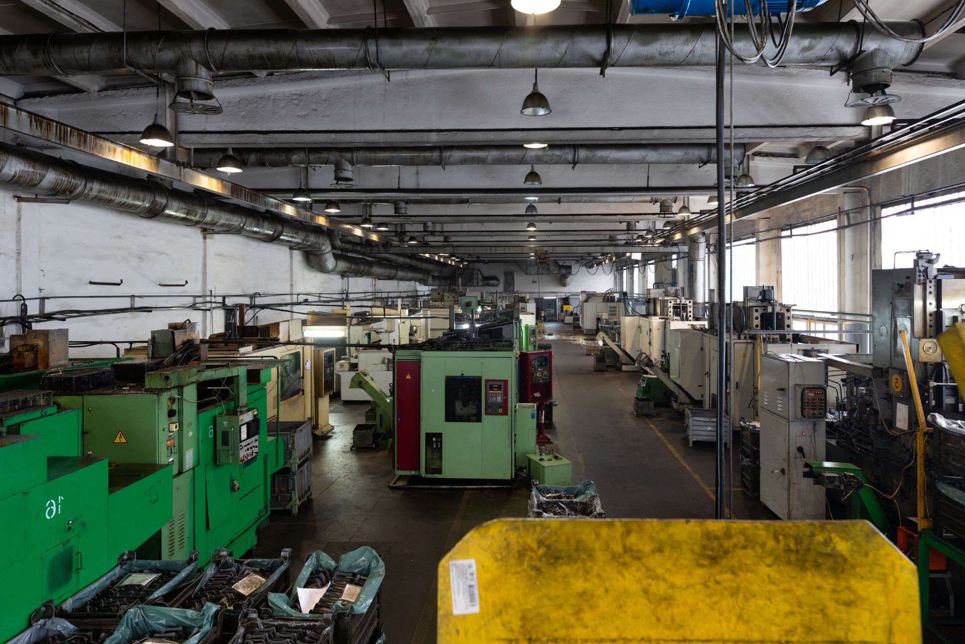 Wide view of an industrial workshop with green machines, overhead pipes, and a yellow bin