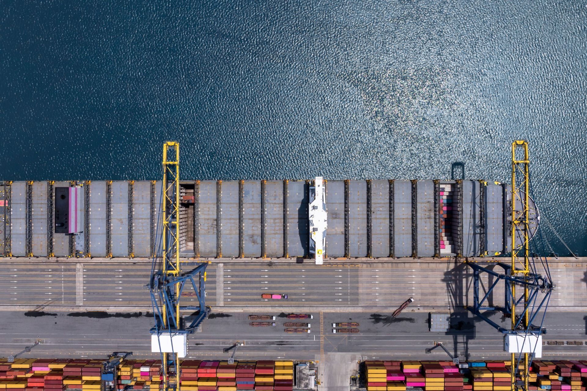 Aerial view of cargo ships docked at a port beside a blue waterfront and stacked shipping containers