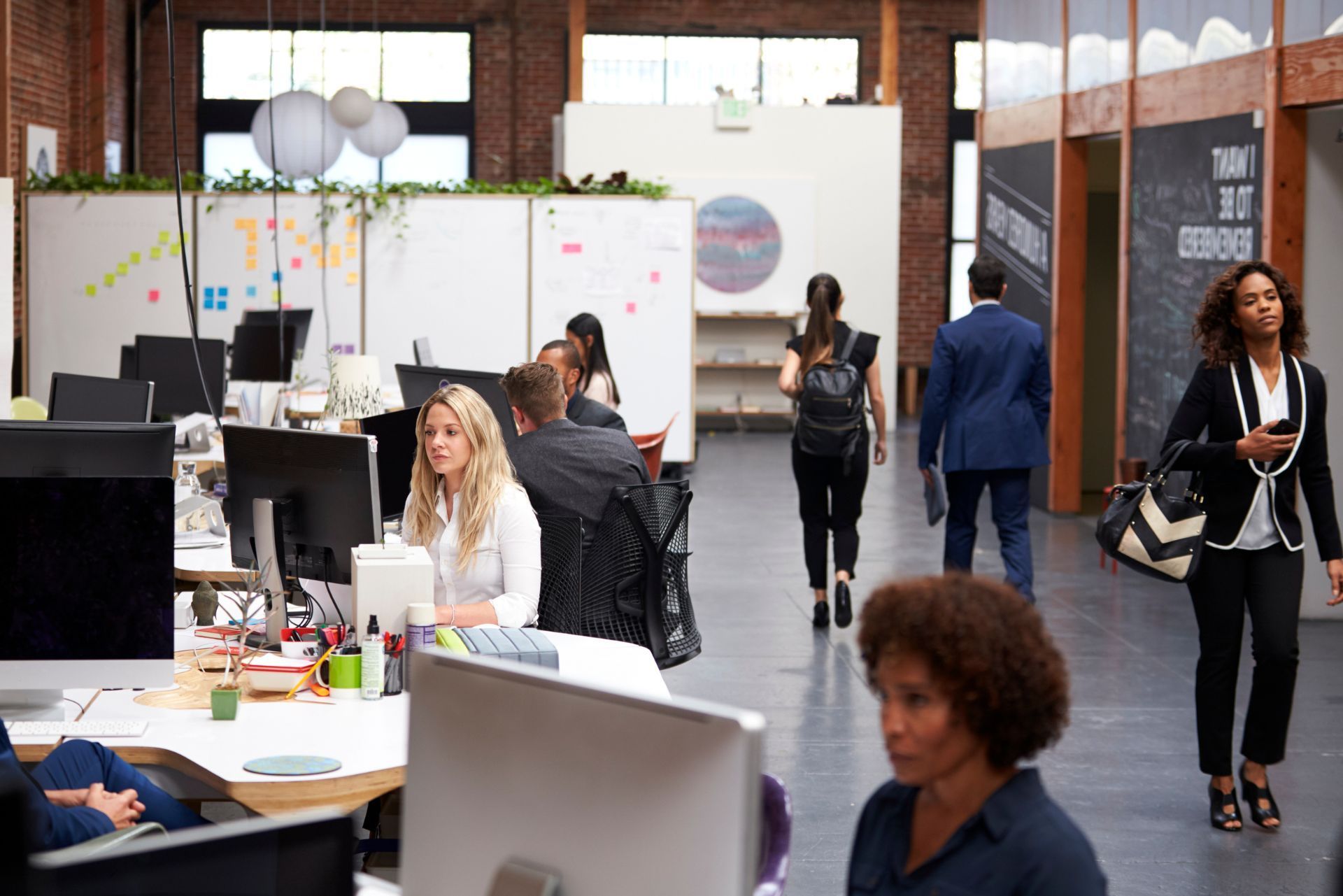 Open office with people working at desks and whiteboards, brick walls and large windows in the background