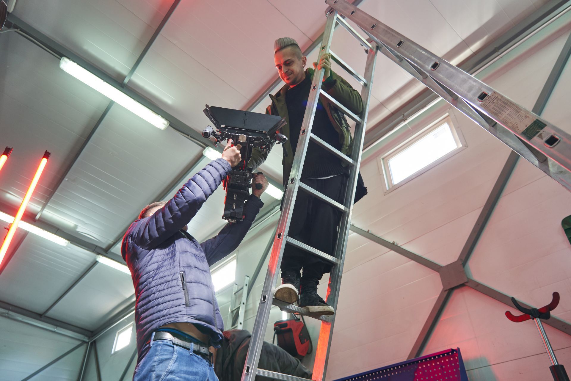 Two workers on a ladder installing equipment on a ceiling indoors
