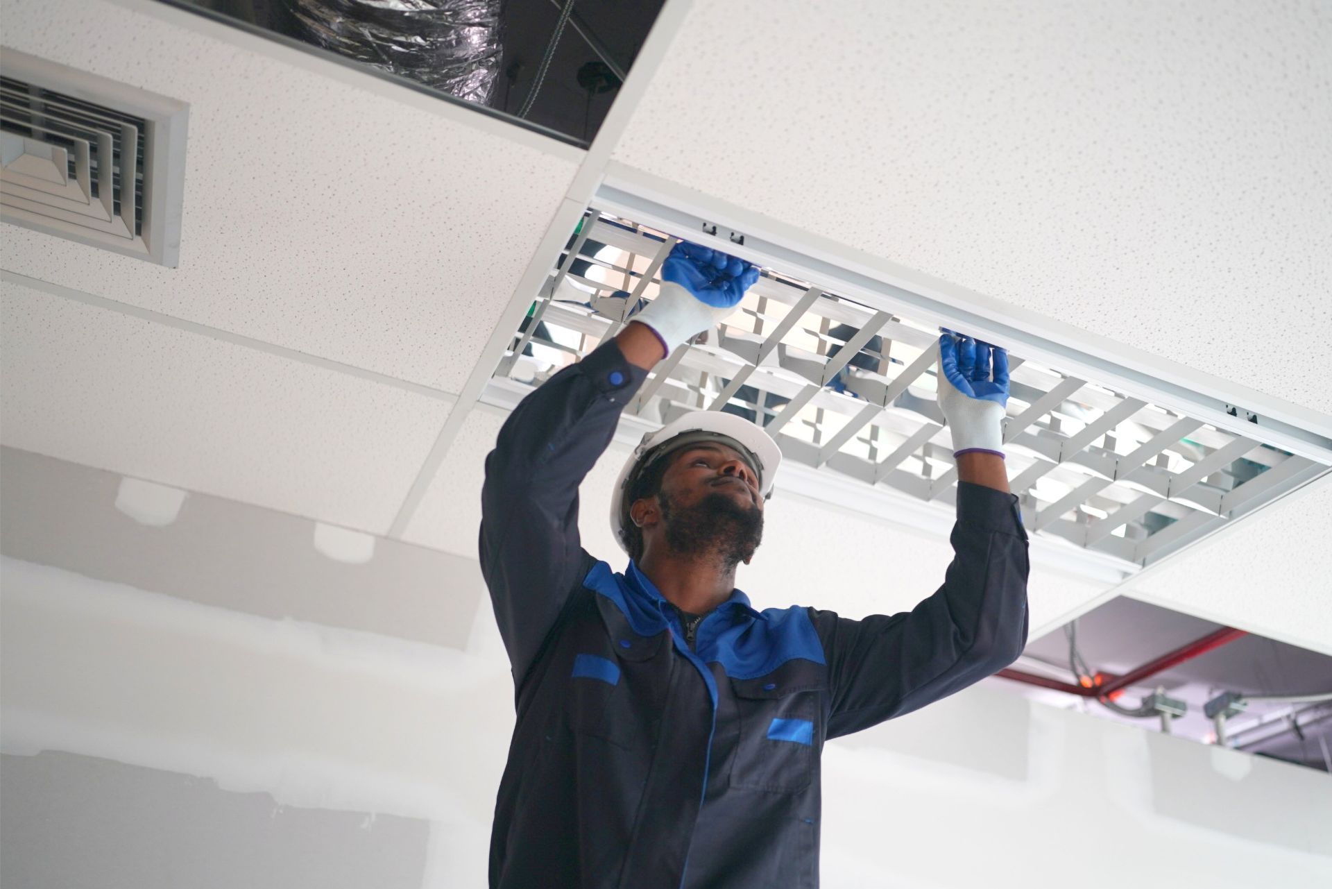 Worker installing a ceiling light panel in a white room, wearing blue gloves and a dark uniform.