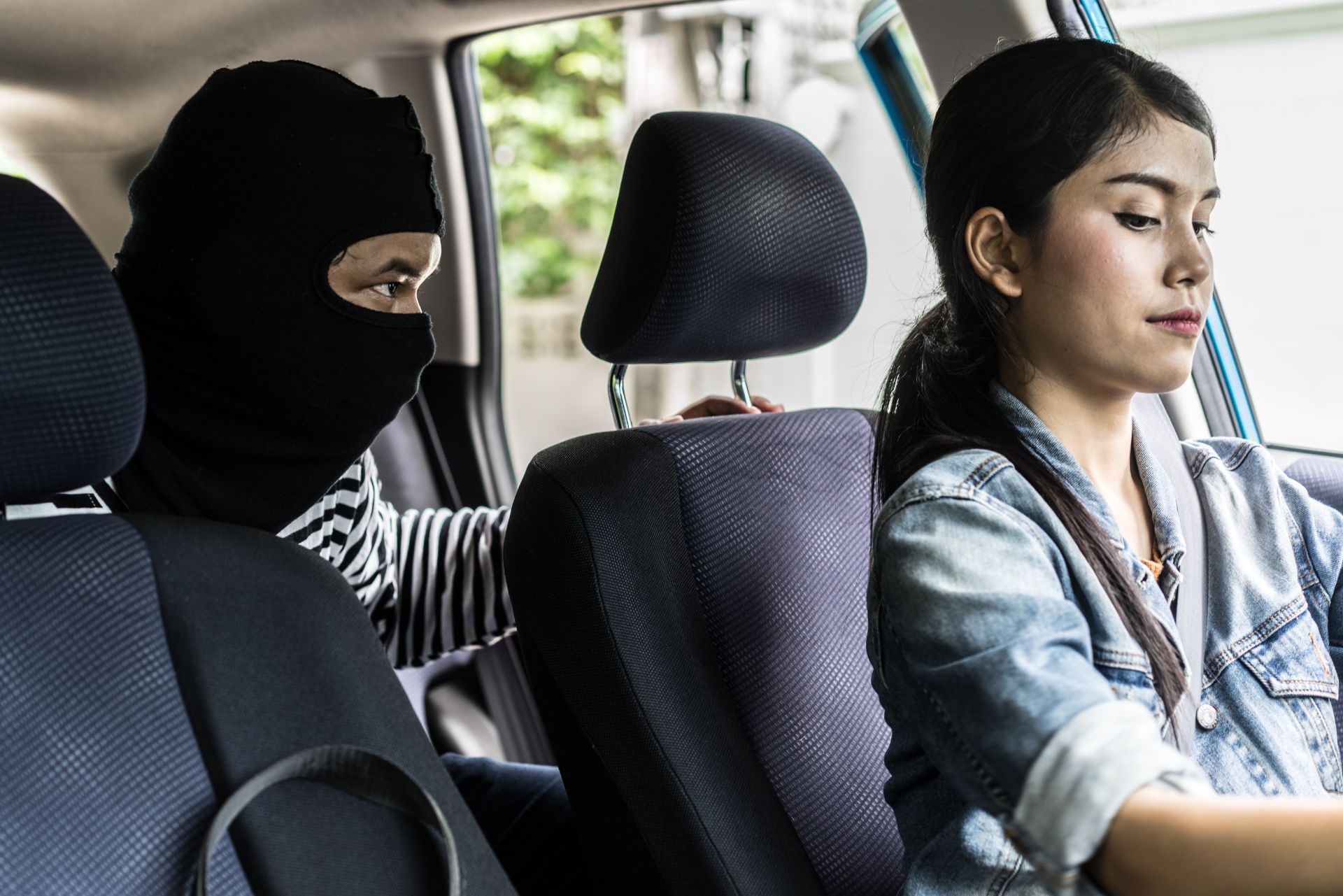 Masked passenger sits in back of a car while a woman drives, both viewed from the front seat area.