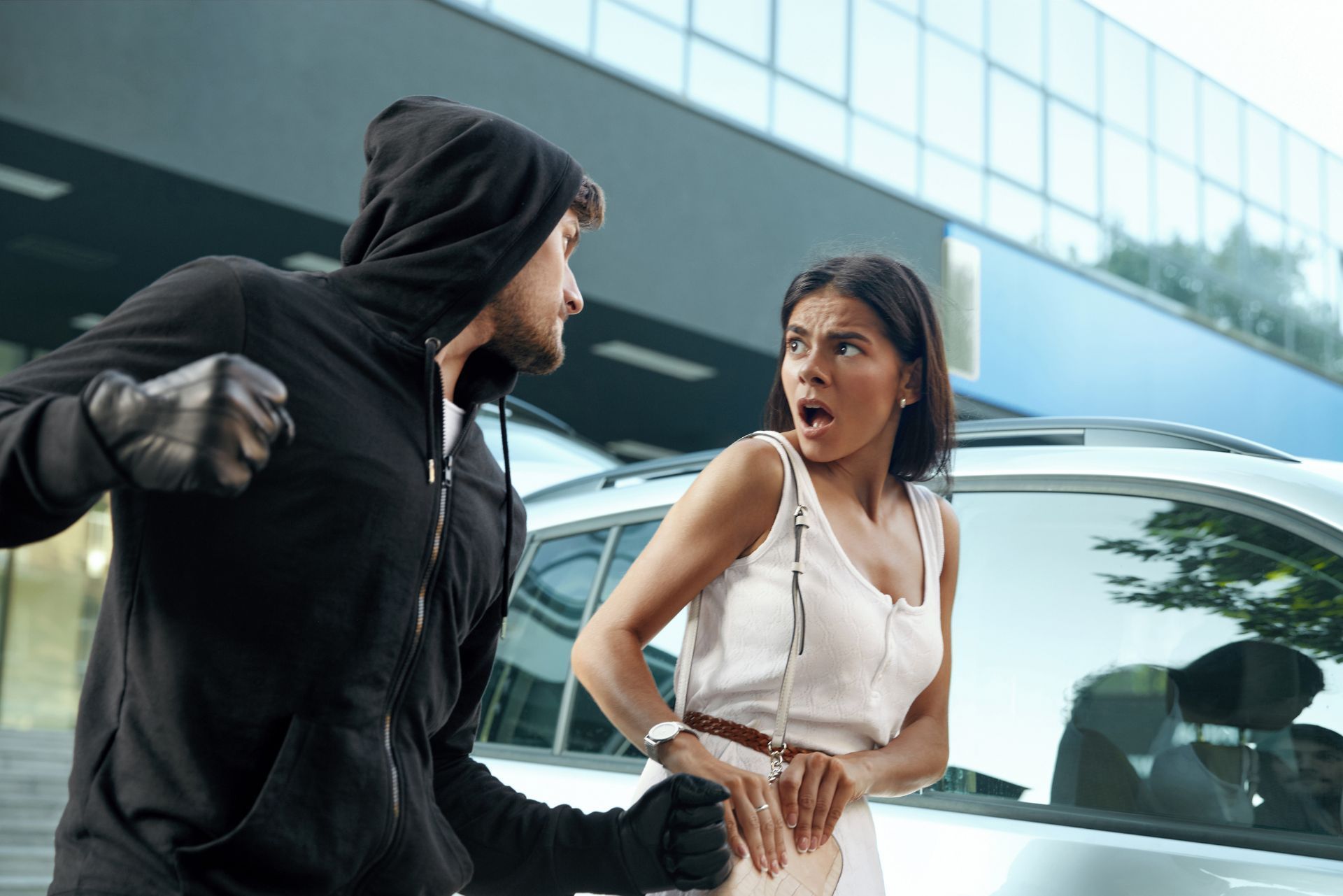 Masked figure confronts startled woman beside a silver car outside a glass building