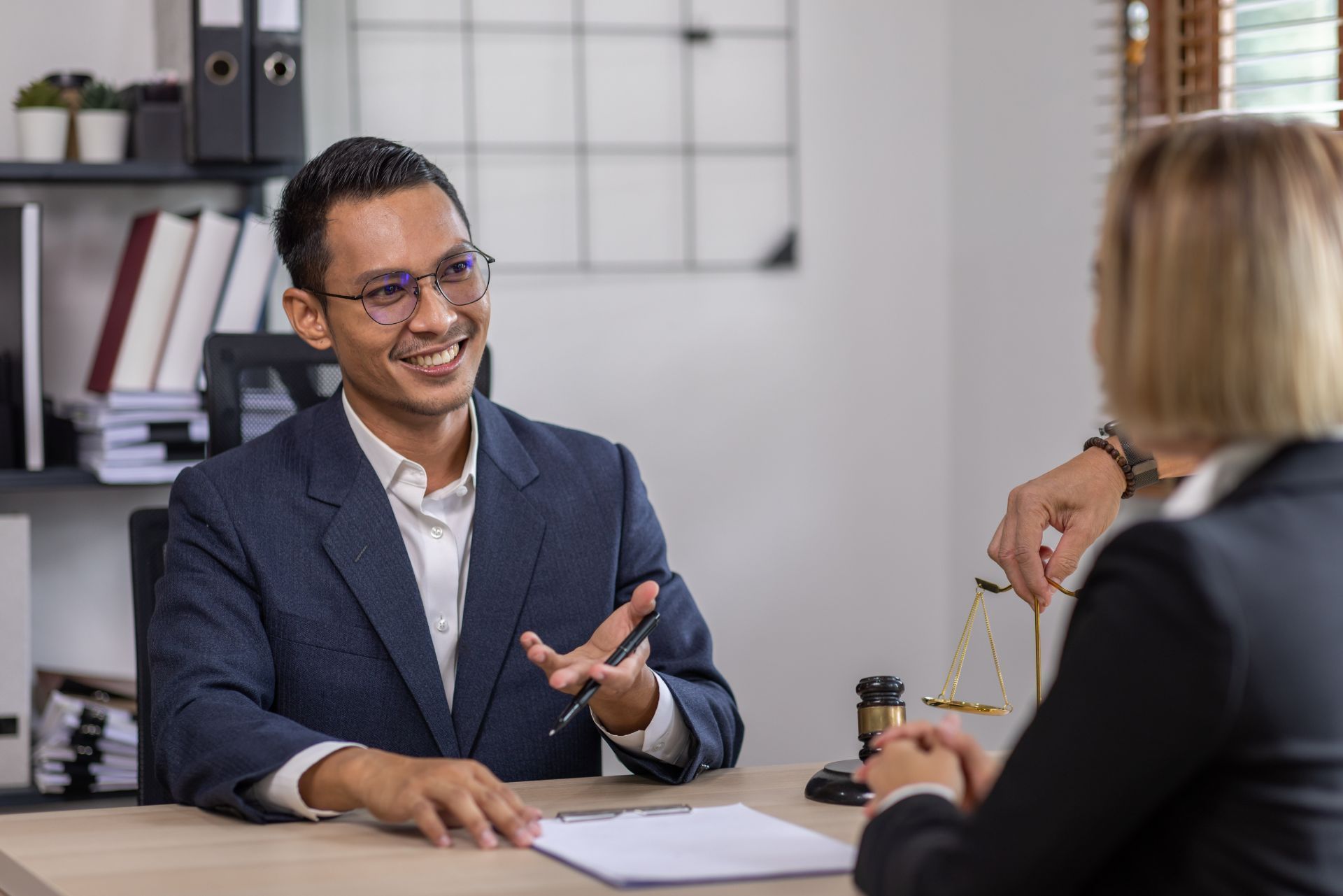 A smiling professional in a suit talks to a client at a desk in a law office with a gavel and balance scales nearby.