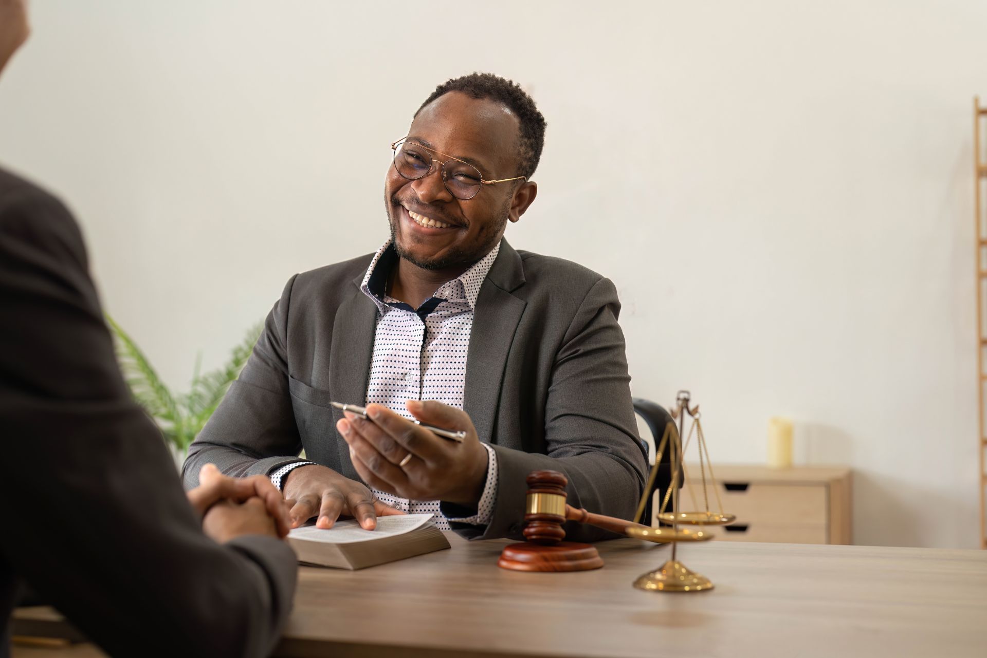 A smiling legal professional in a suit sits at a desk with a gavel and balance scale, facing a client.