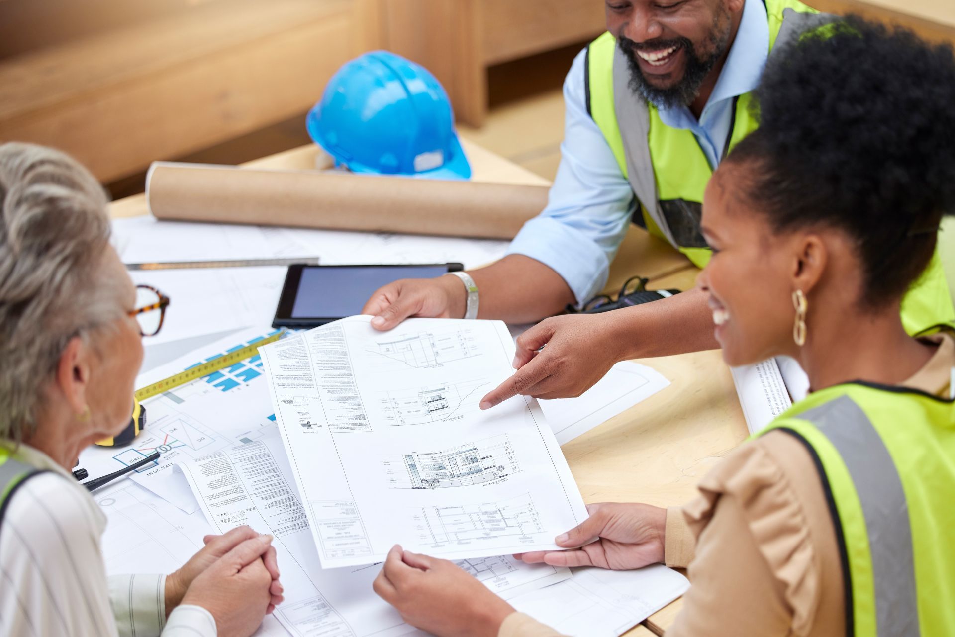 Three coworkers in safety vests reviewing blueprints at a construction site table