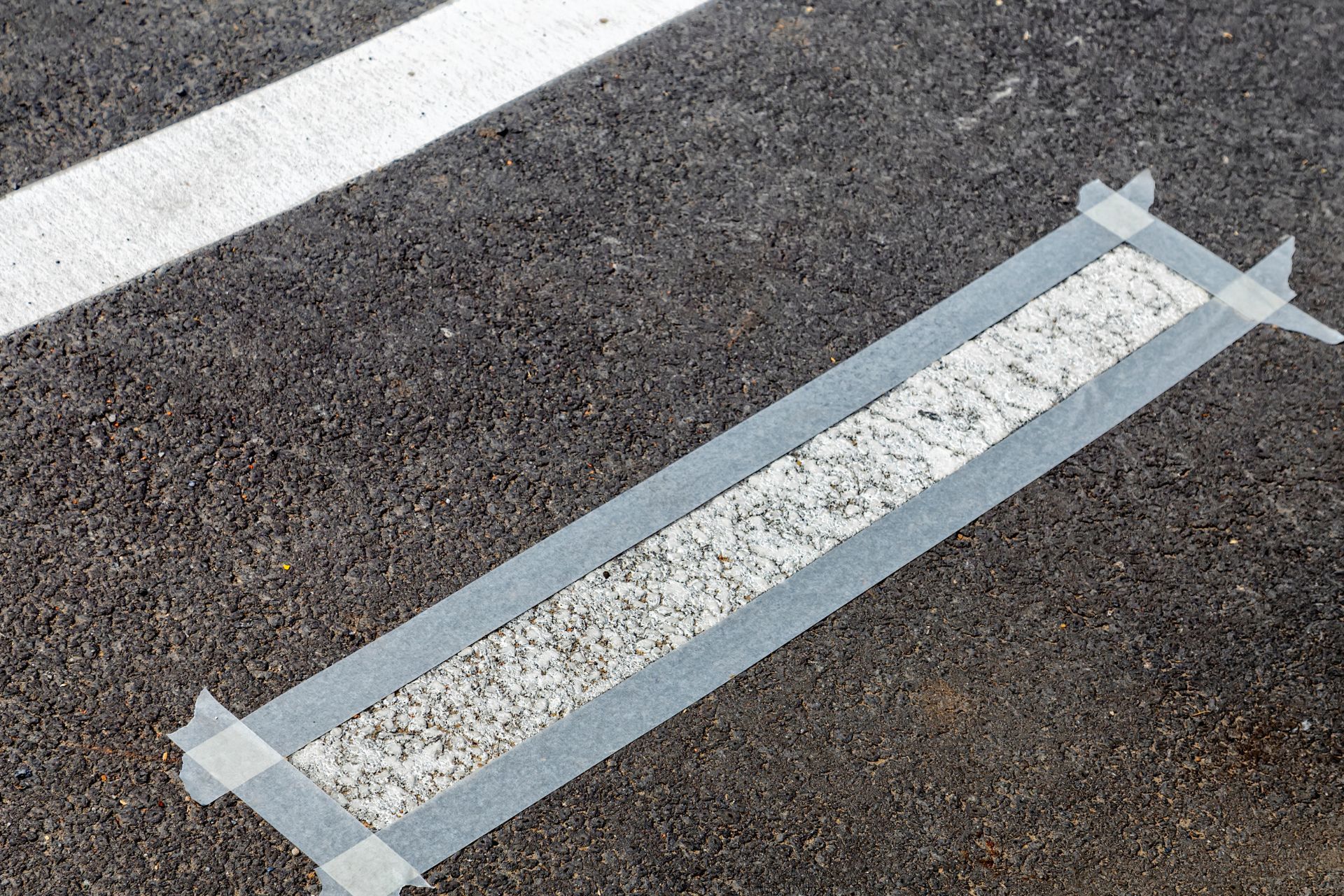 A rectangular section of asphalt is framed with masking tape, creating a border around a light-colored, pebbled strip.