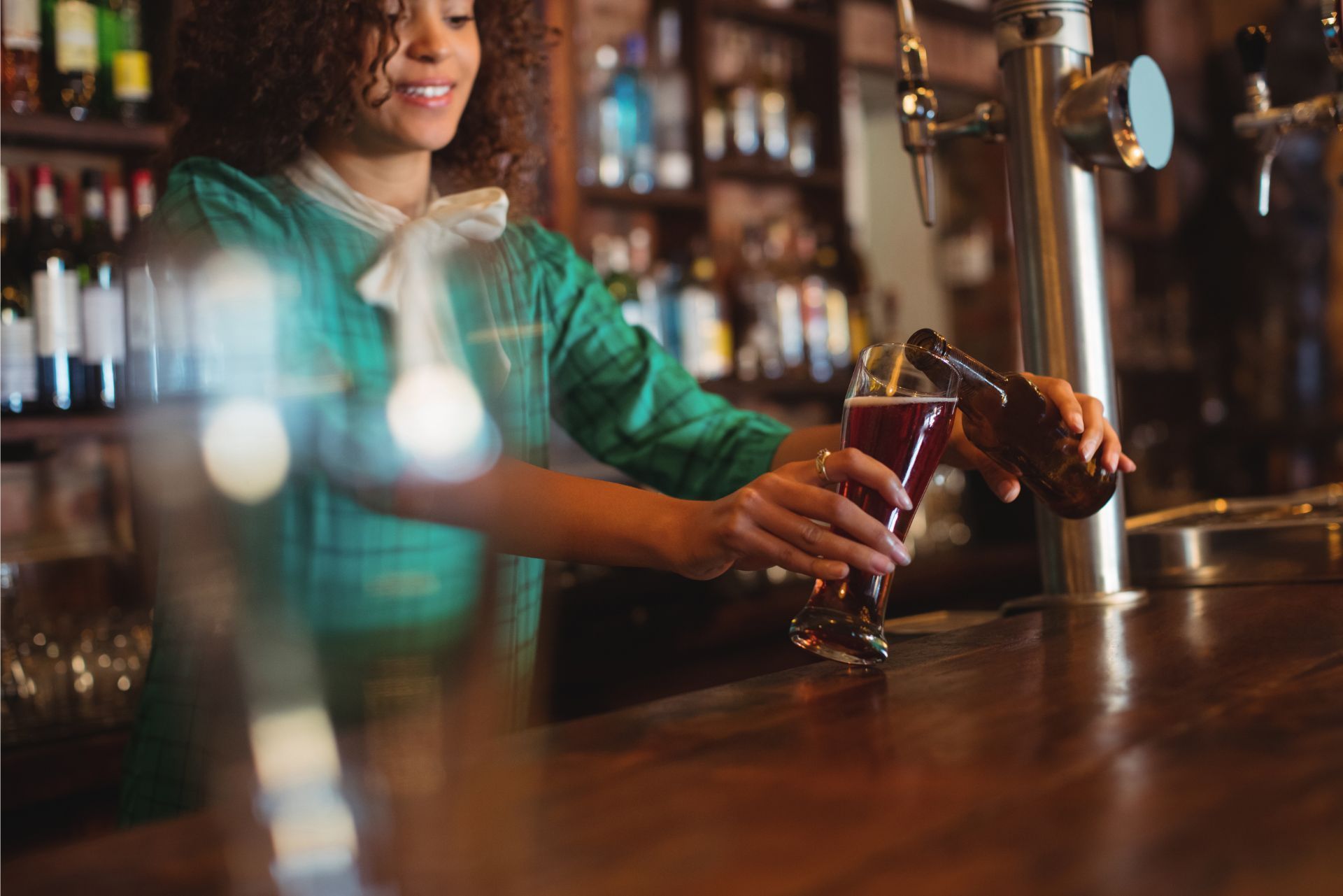 Bartender pouring a drink at a wooden bar, with bottles and glassware blurred in the background.