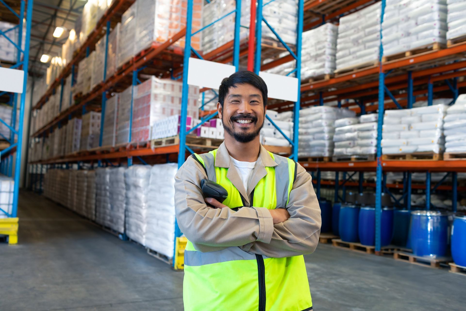 Warehouse worker in a neon vest stands with arms crossed in an aisle of stacked pallets and drums.