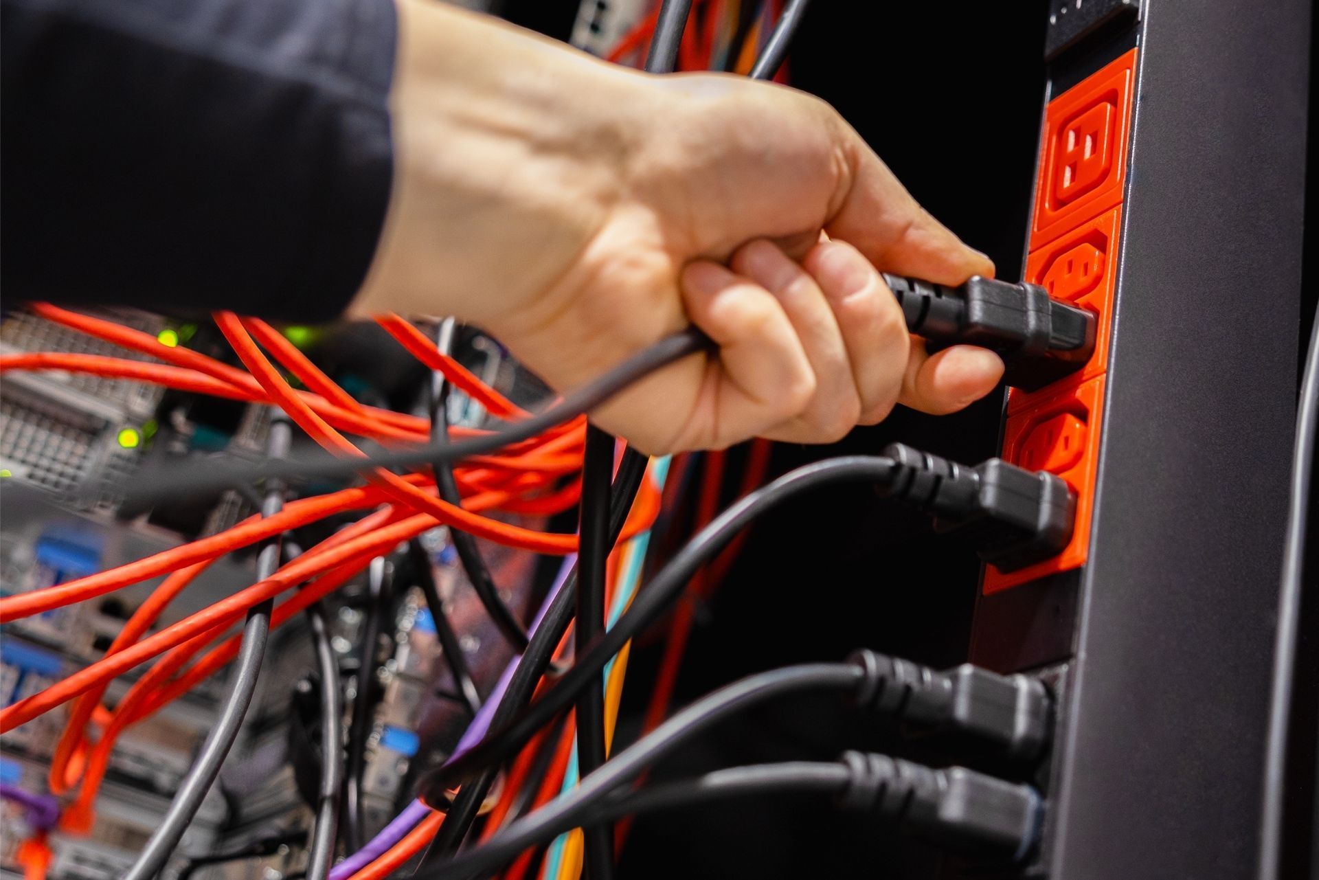 A hand plugs a black power cable into a red power strip inside a server rack with many tangled red and black cables.
