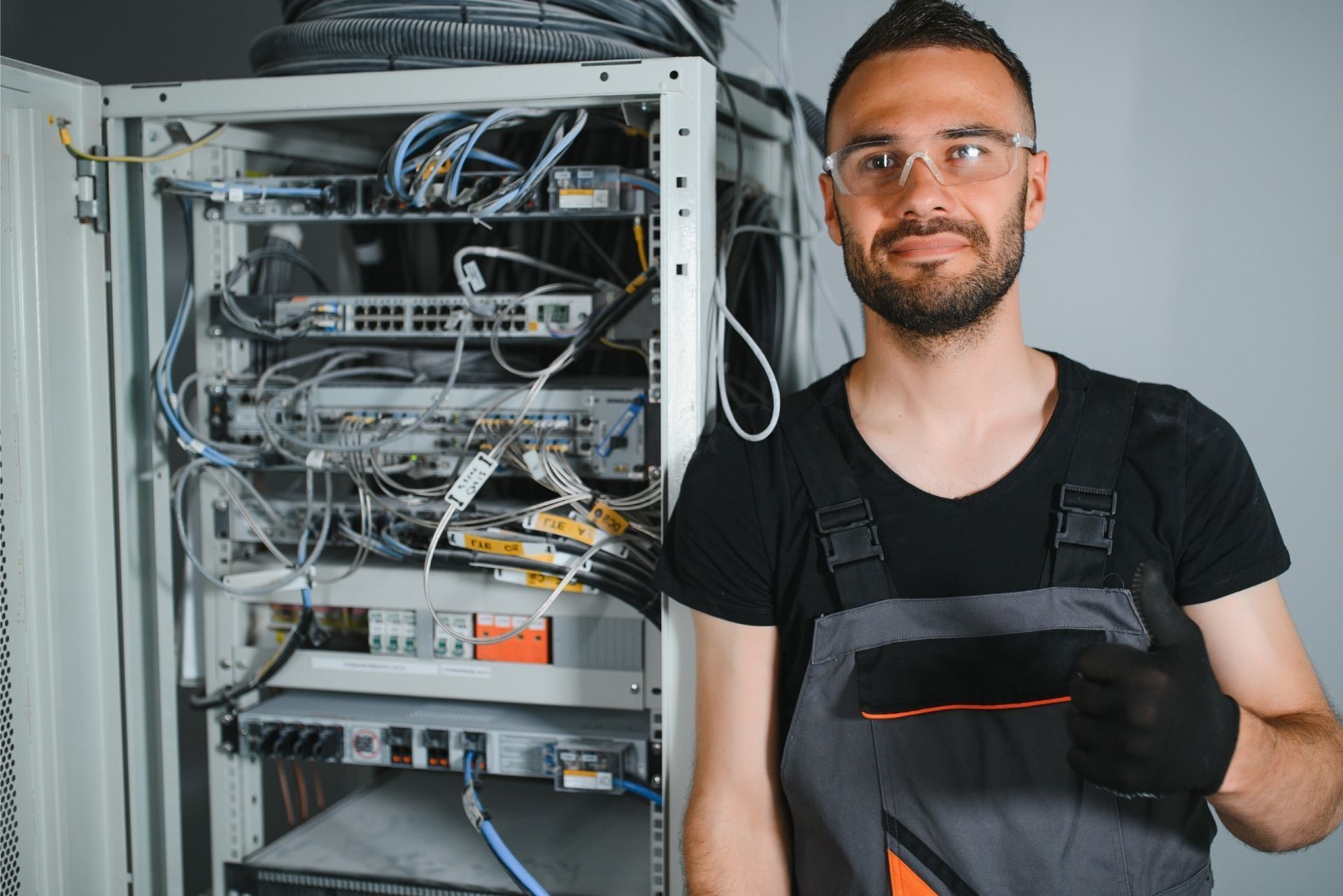 A technician wearing safety glasses and work coveralls stands next to an open server rack with networking equipment.