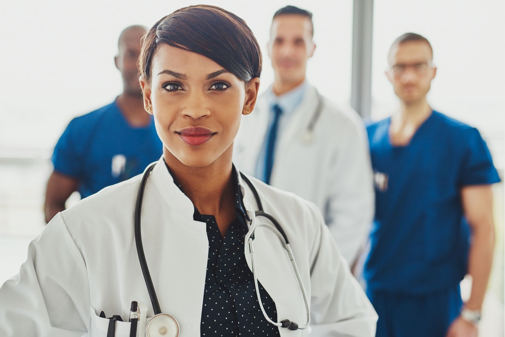 A medical professional in a white coat and stethoscope stands in front of colleagues in a bright, professional setting.