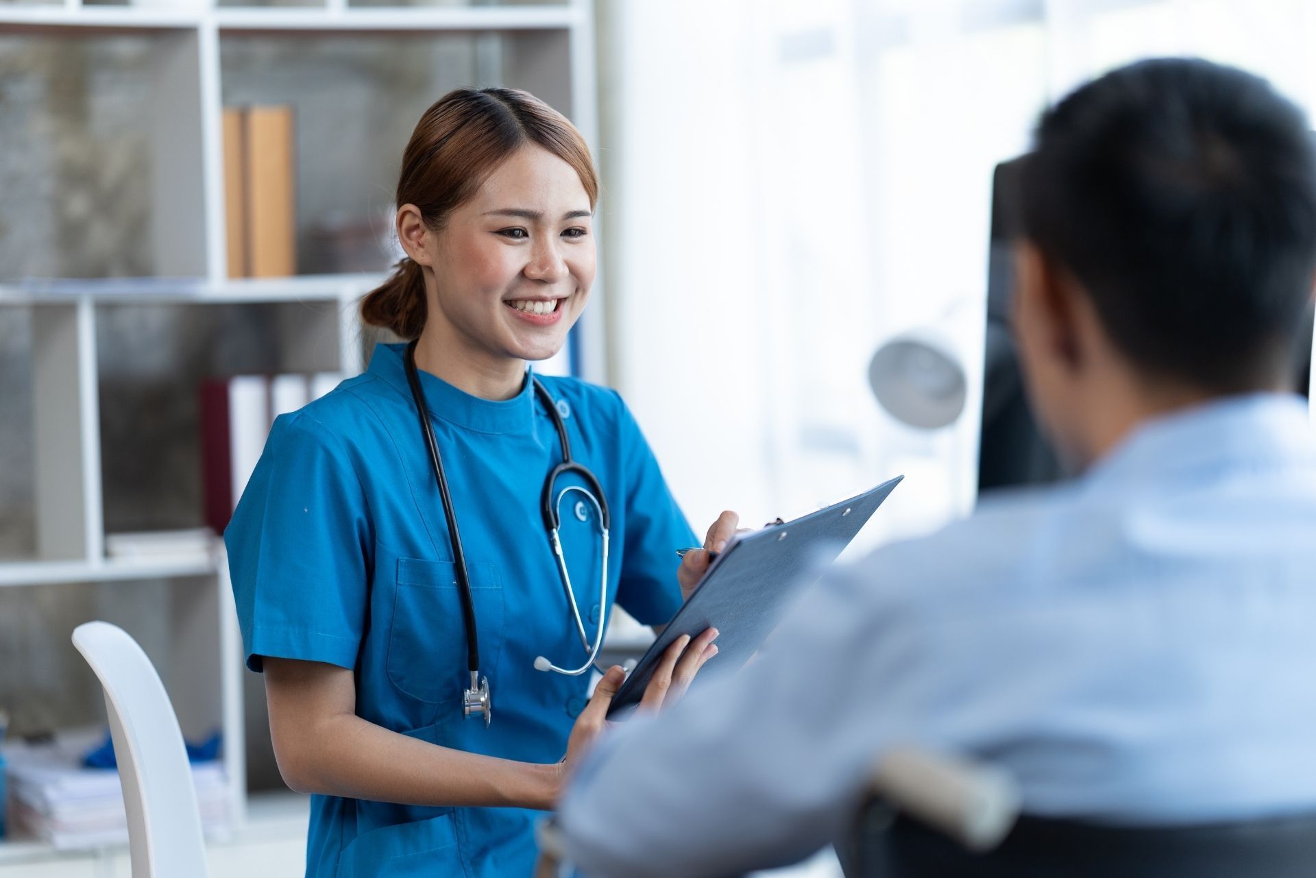 A smiling healthcare worker in a blue uniform holds a clipboard while speaking with a patient in an office.