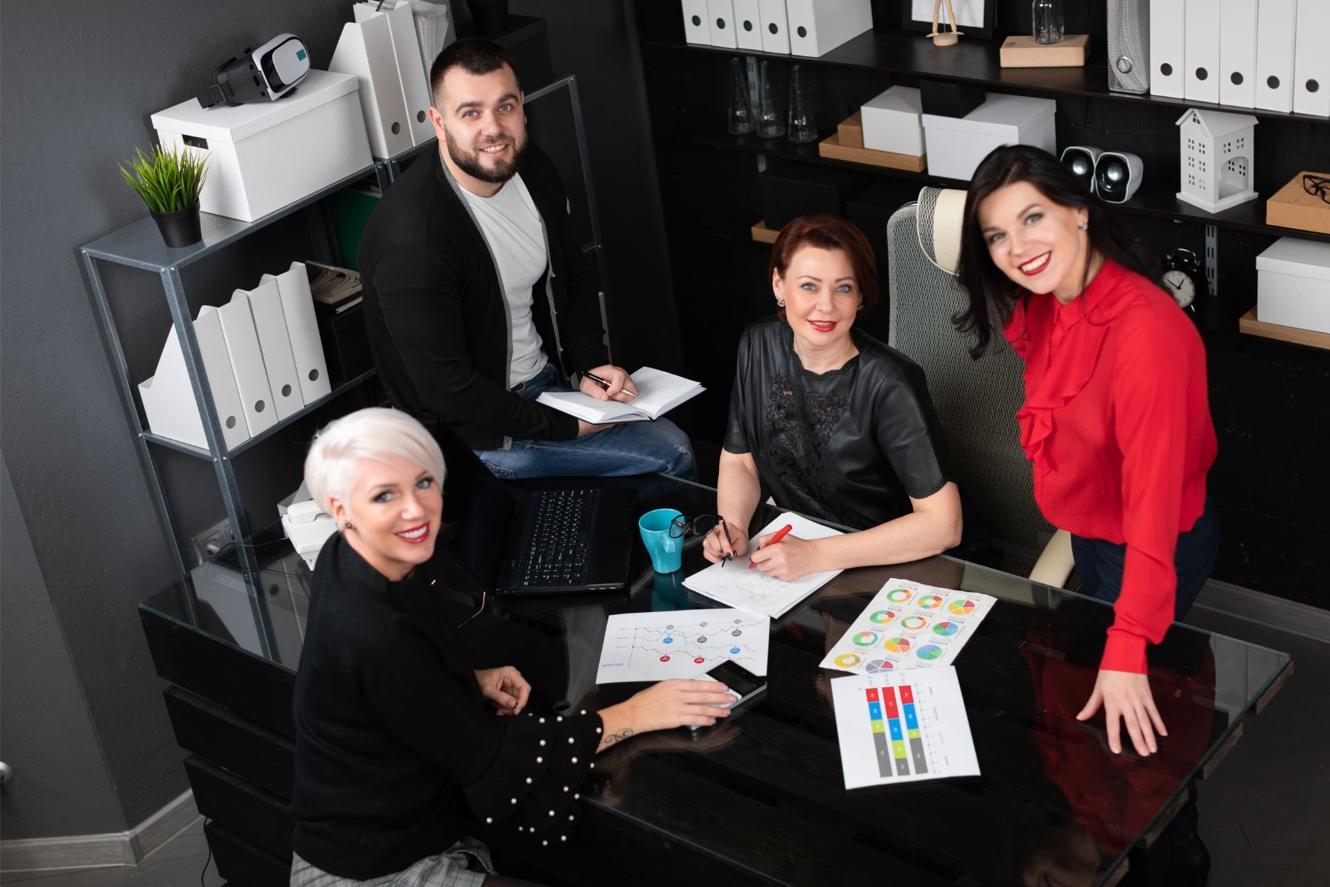 Four coworkers in a modern office review documents around a desk, smiling at the camera.