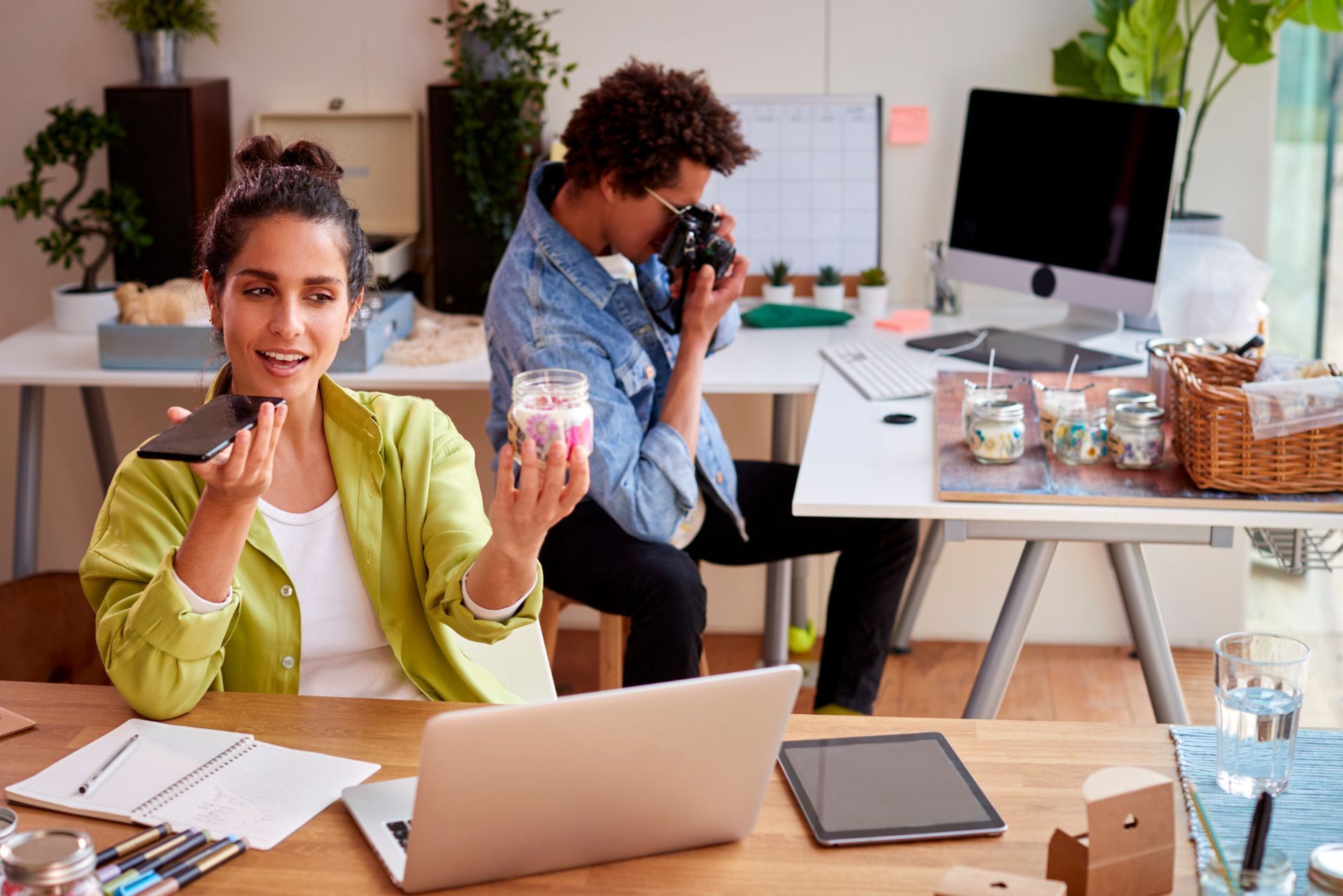 Two coworkers in a bright office, one speaking on the phone and holding a coffee cup while the other takes a photo.