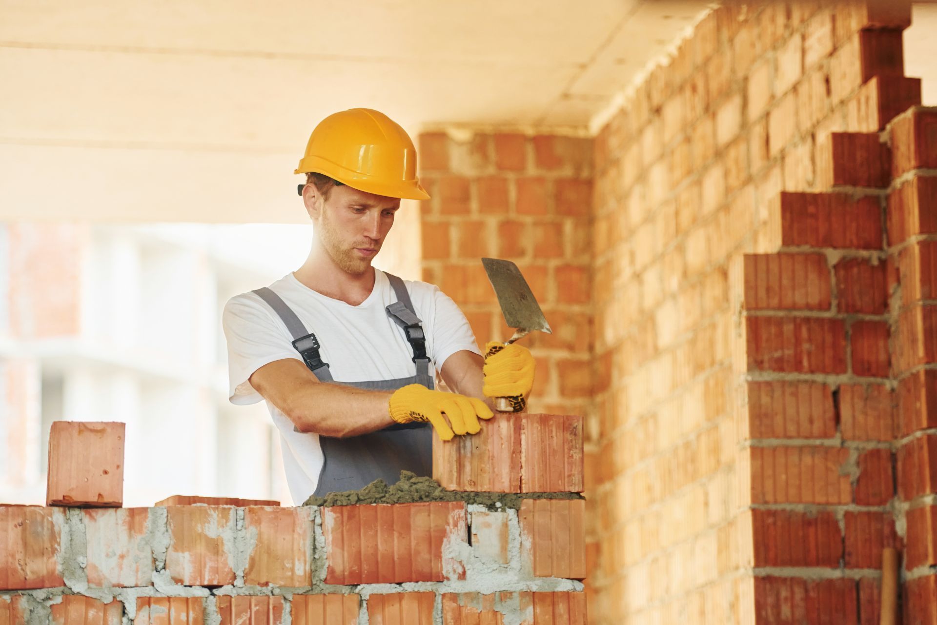 A construction worker in a hard hat and safety gloves lays a brick with a trowel on a masonry wall.